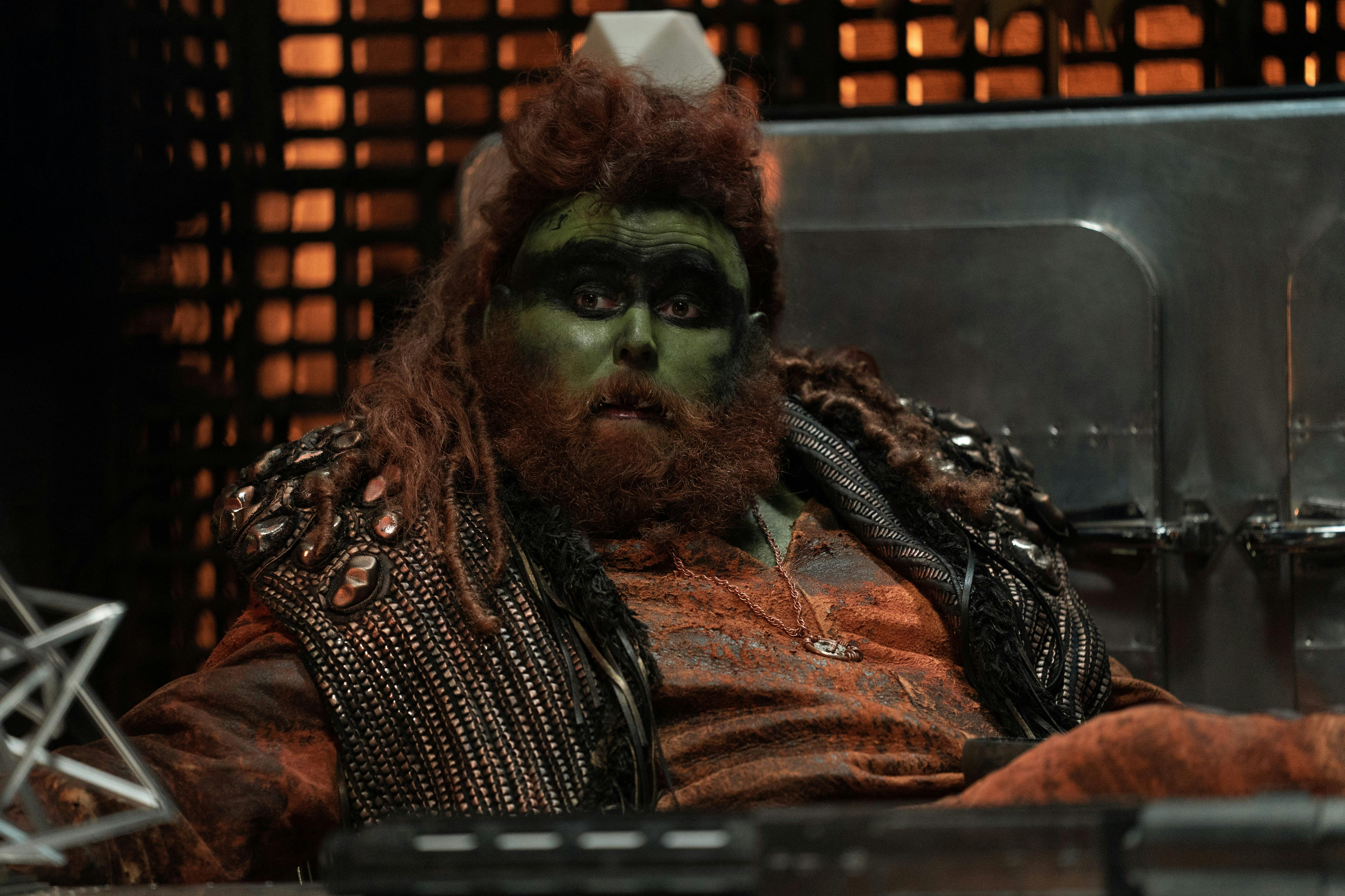 A green alien with red hair and a red beard looks quizzically at someone off camera. He is sitting behind a desk.