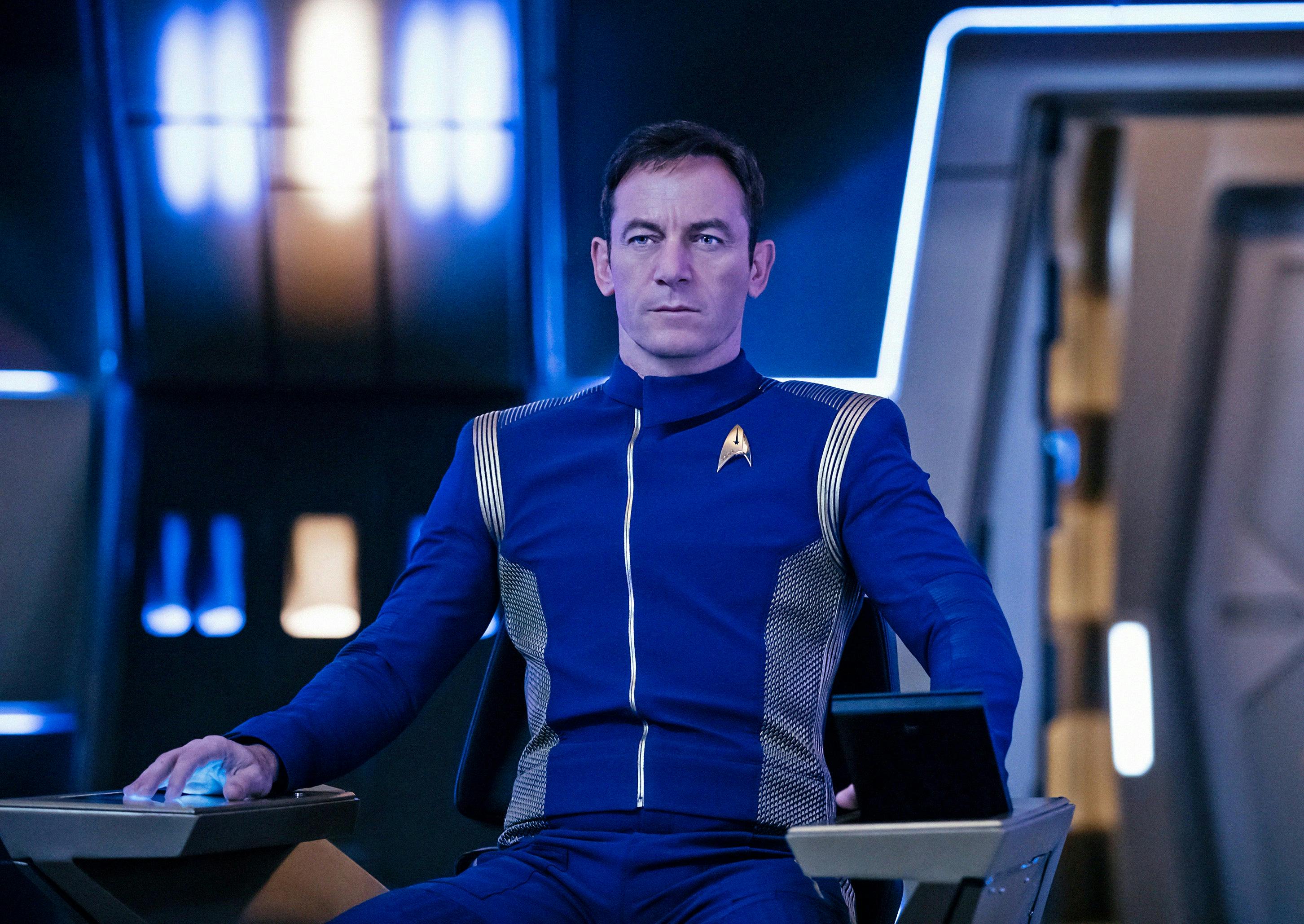Lorca (Jason Isaacs), wearing the blue Discovery uniform, sits in the captain's chair on the bridge.