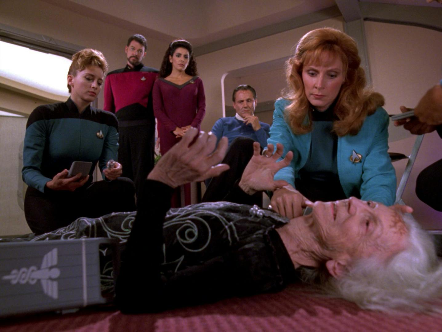 Crouching on the ground, Dr. Crusher investigates the suspicious death of a Lumerian ambassador's mother in 'Man of the People'