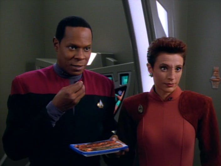 Ben Sisko raises a snack to eat as he stands next to Kira Nerys in 'Sanctuary'