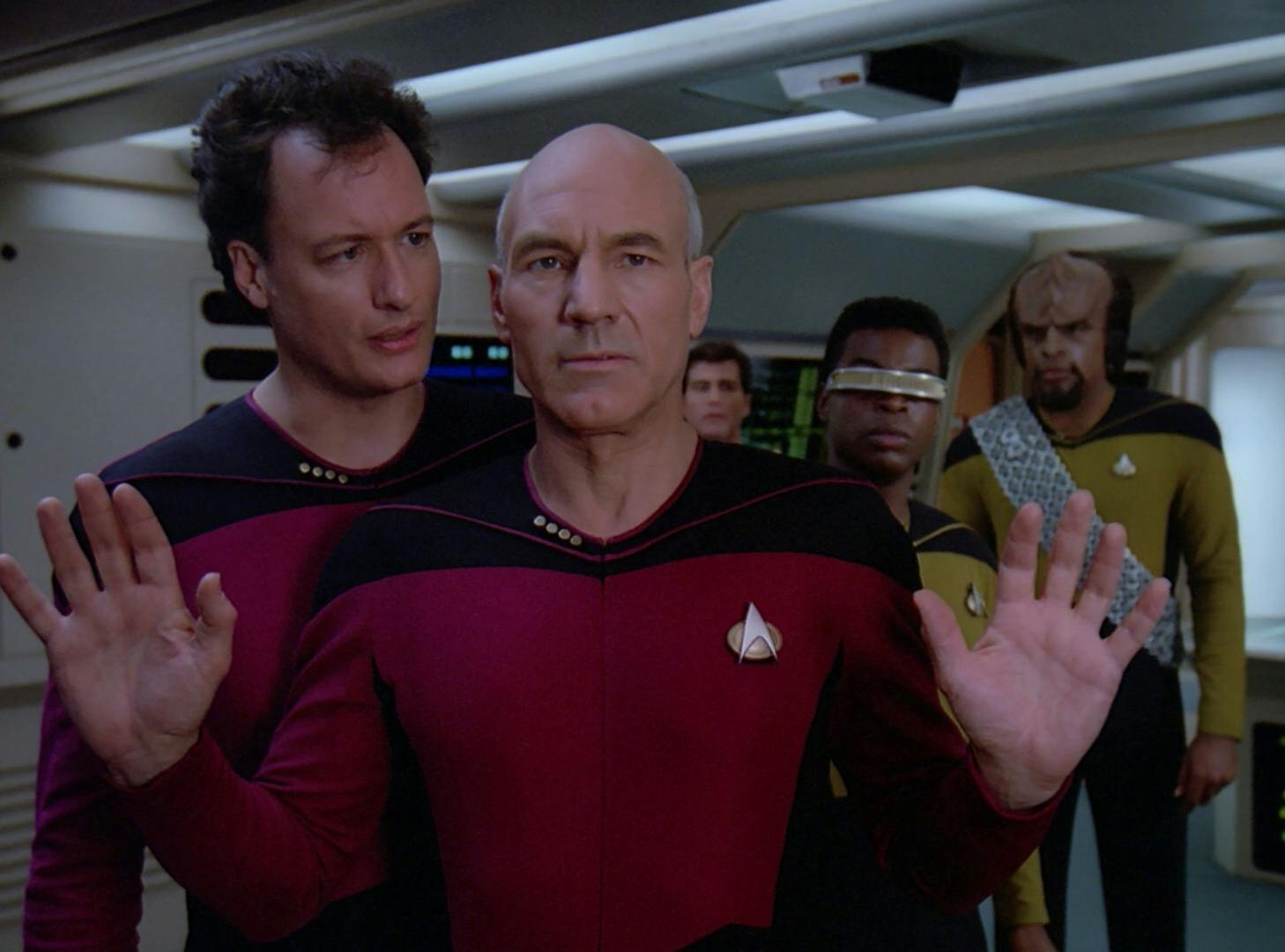 Picard concedes with both hands raised up in defeat as Q, Geordi La Forge, and Worf stand behind him in 'Q Who'