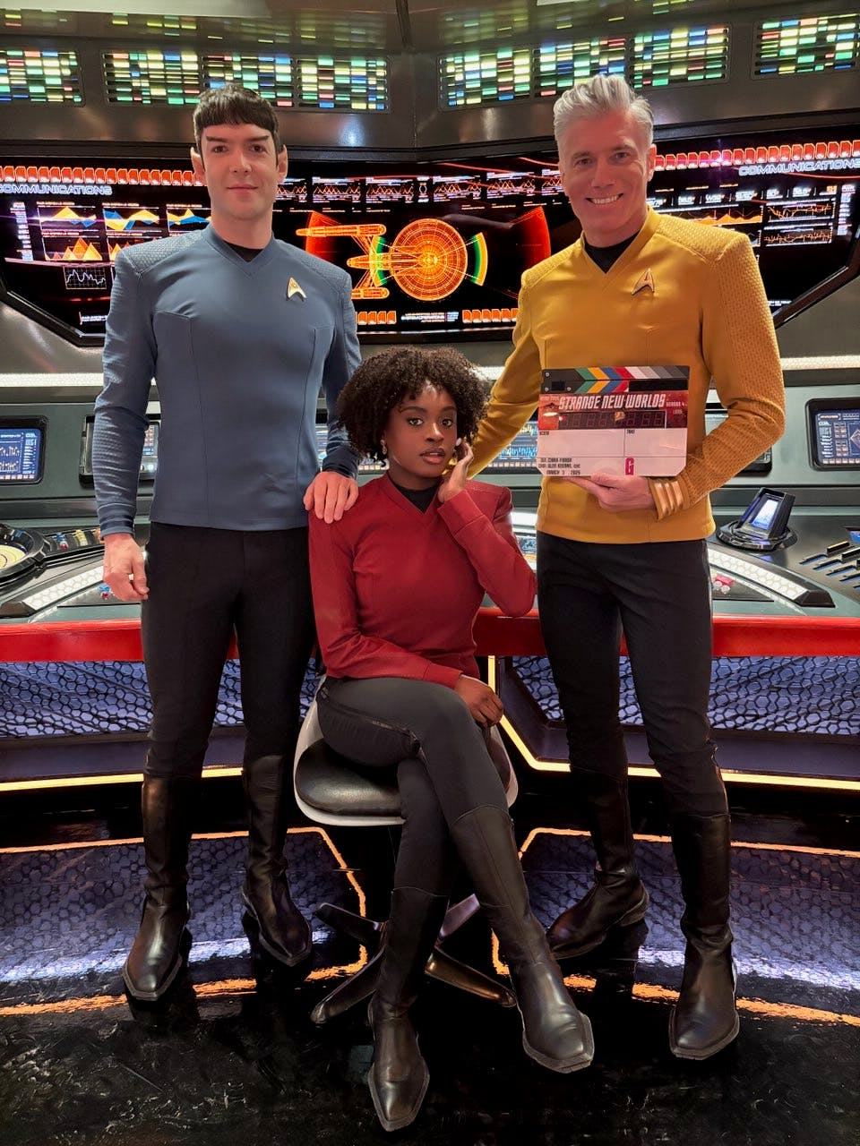 Ethan Peck (Spock), Celia Rose Gooding (Uhura), and Anson Mount (Christopher Pike) holding a clapboard pose for a photo on the bridge set of Star Trek: Strange New Worlds