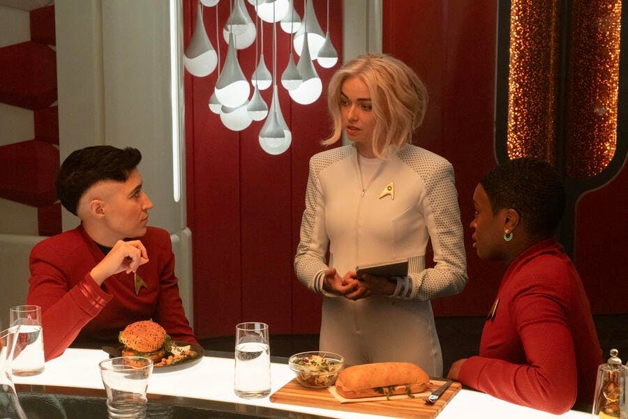 Christine Chapel holding a PADD approaches the seated Erica Ortegas and Uhura having dinner in the mess hall in 'Charades'
