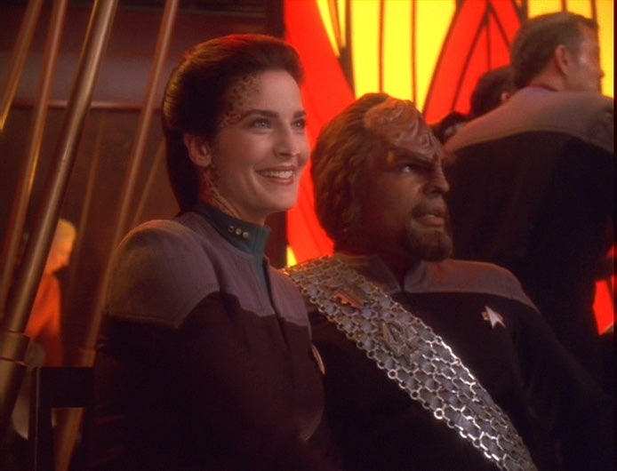 Jadzia Dax and Worf sit side-by-side at Quark's as she smiles deeply in 'You Are Cordially Invited'