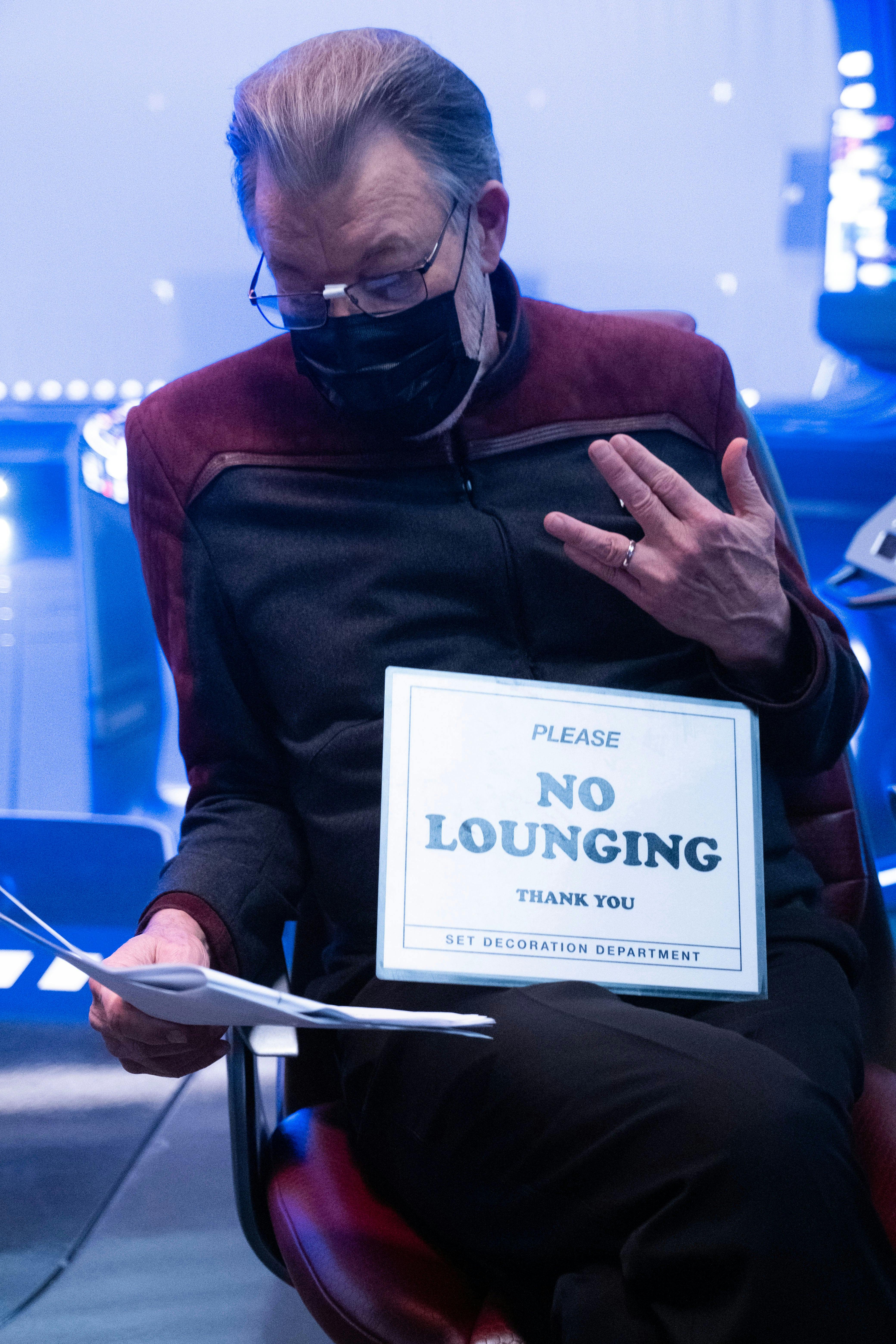 Star Trek: Picard BTS still - Jonathan Frakes reading his script displaying a Vulcan salute with a 'No Lounging' sign on his lap