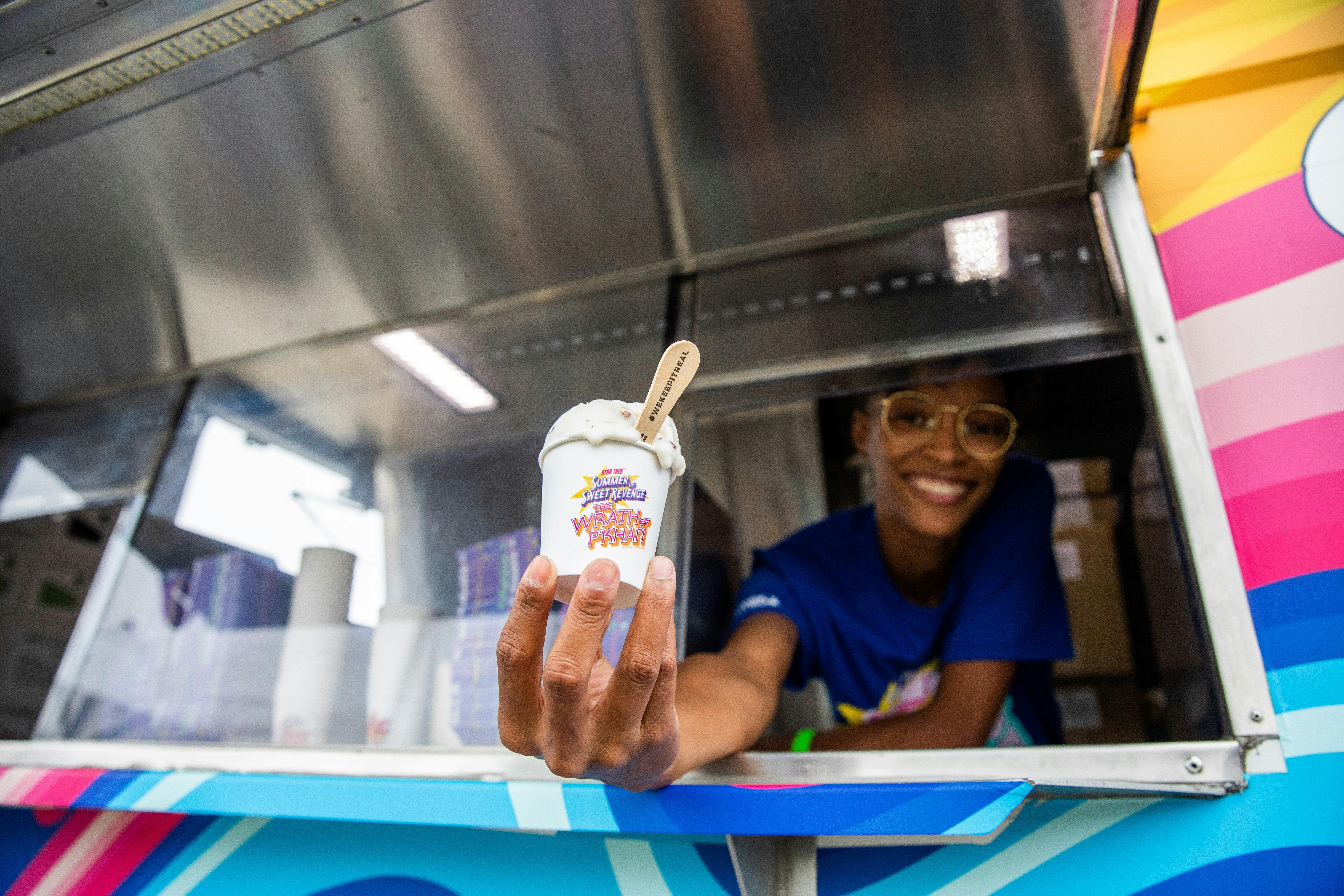 A smiling worker holds an ice cream cup towards the camera.