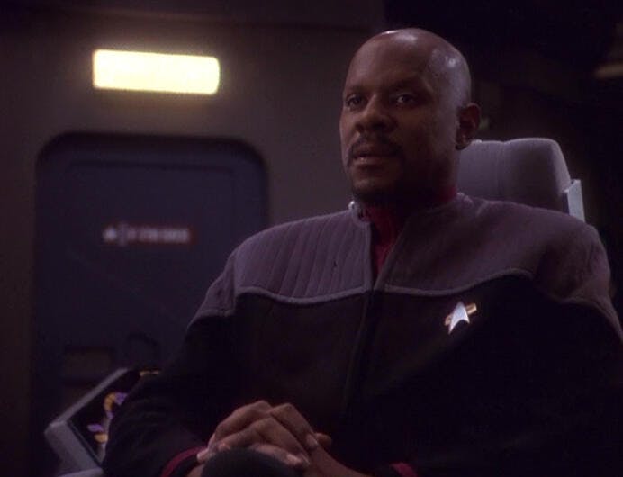Captain Sisko (Avery Brooks) sits on the bridge of the Defiant, hands folded in front of him.