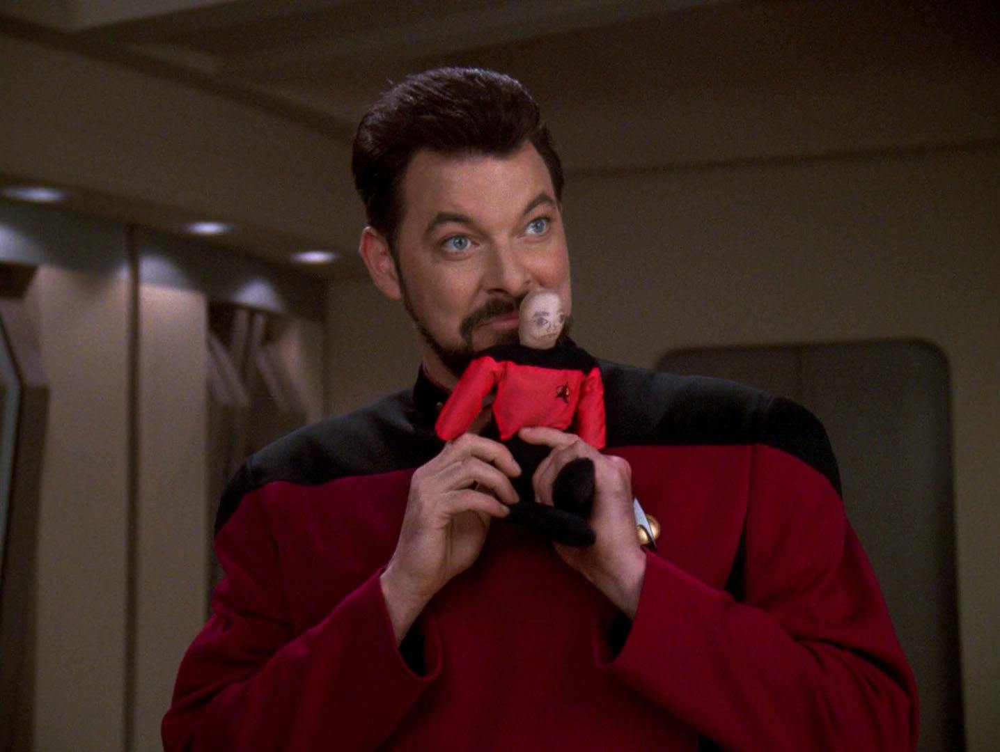 Riker holds a Picard doll to his face while smiling on The Next Generation - The Pegasus