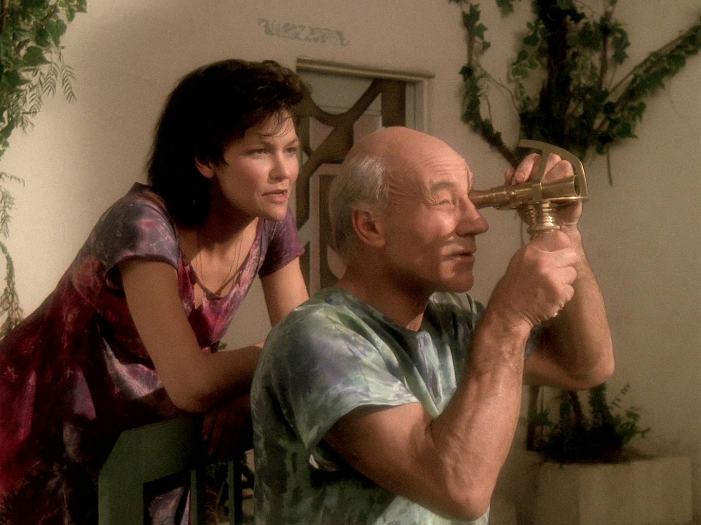 Picard as Kamin in his mindscape looks out through a telescope as his wife leans on the back of his chair behind him in 'The Inner Light'
