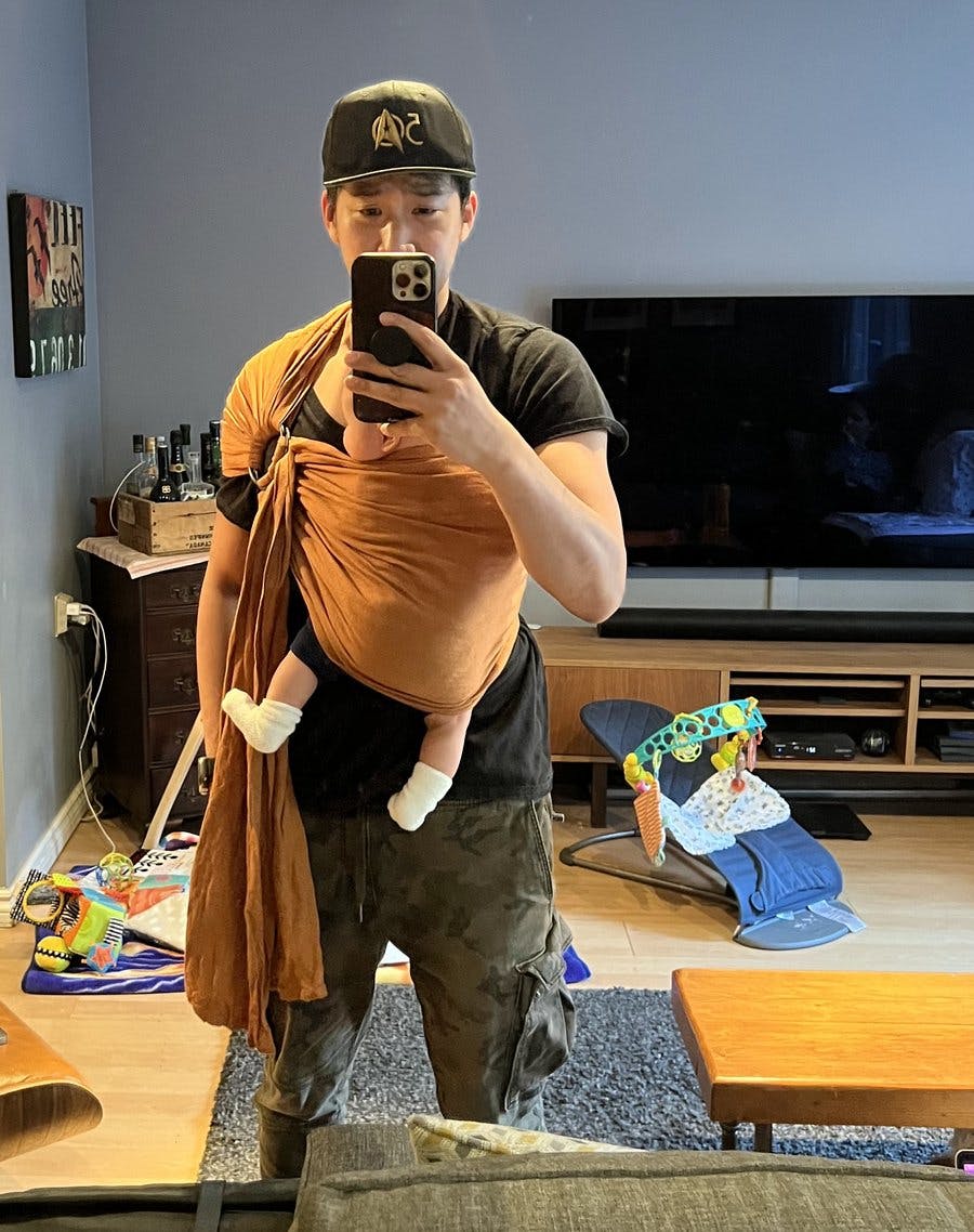 Patrick Kwok-Choon takes a selfie in his living room with his baby swaddled against his body