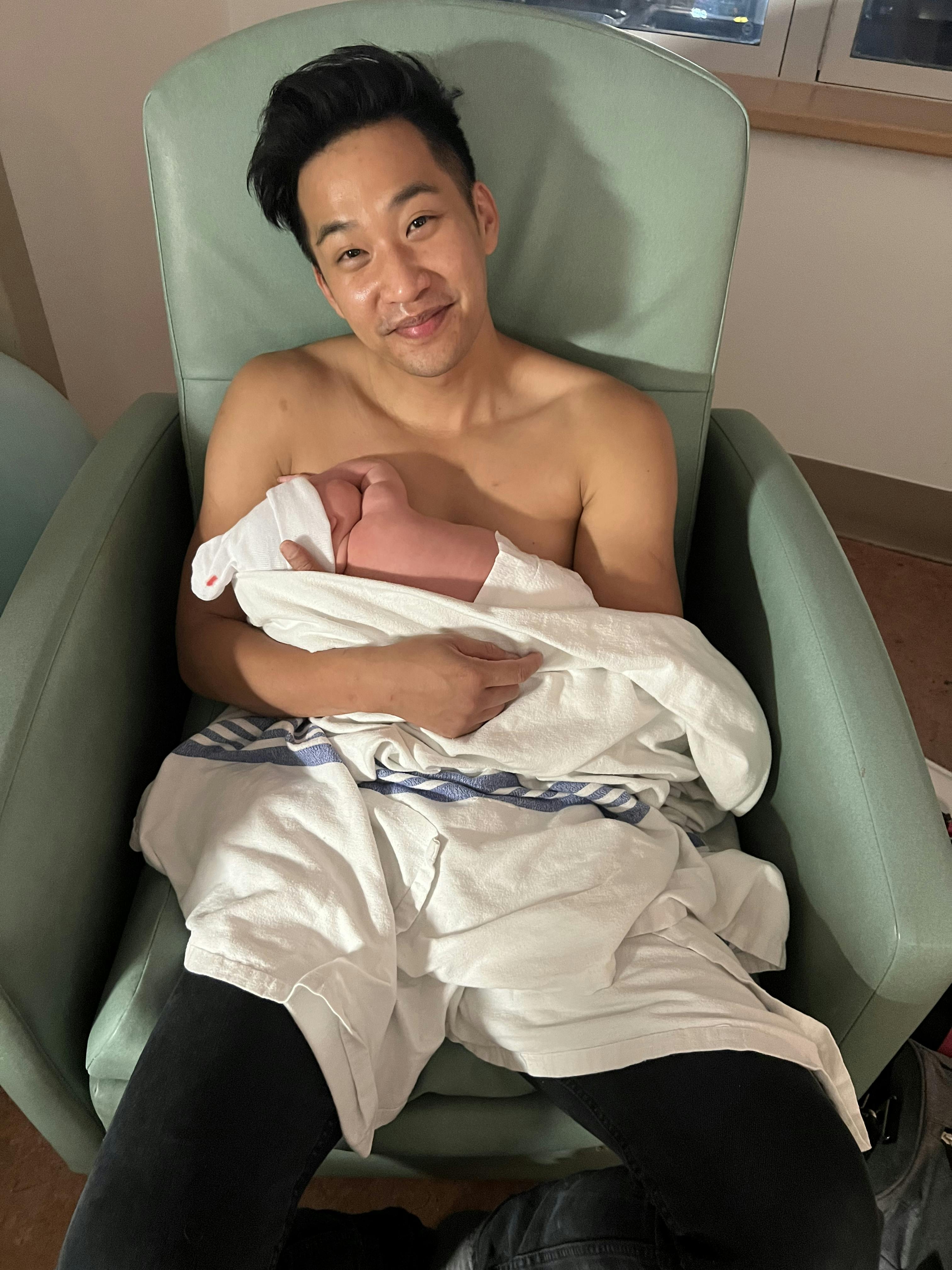 A shirtless Patrick Kwok-Choon holds his newborn baby against his chest