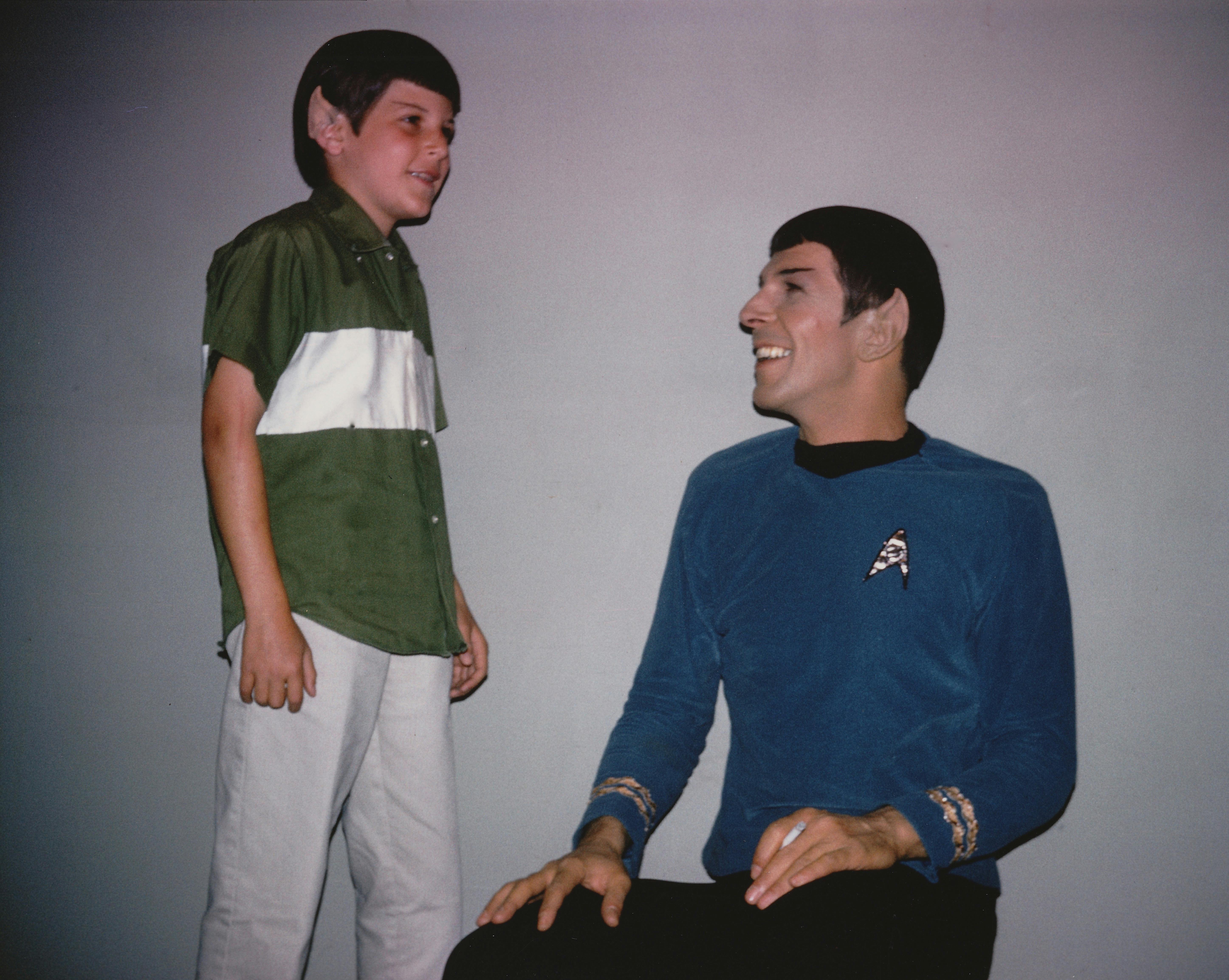 A seated Leonard Nimoy in costume as Spock brightly looks up at his son Adam dressed as his mini Spock