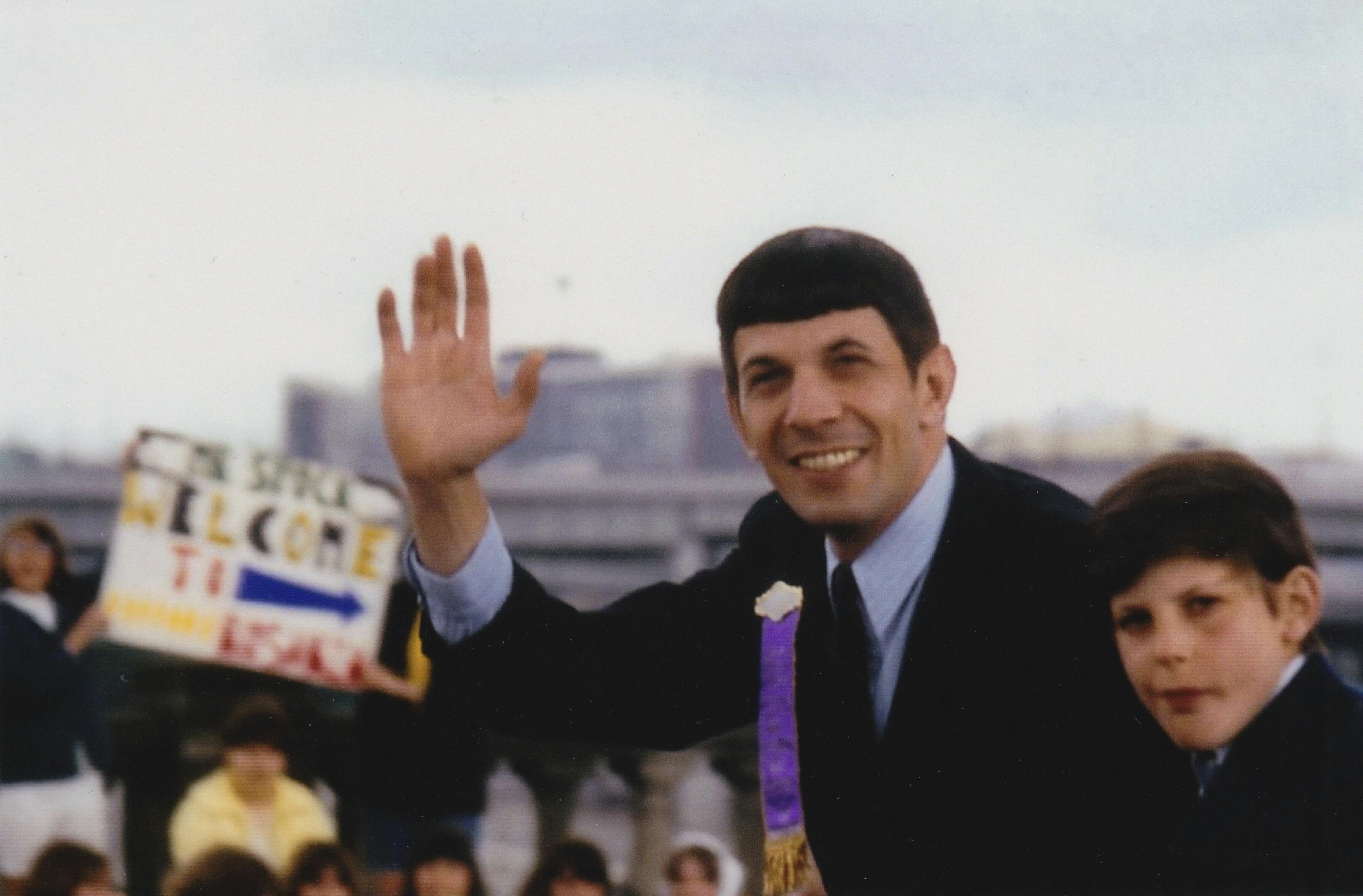 At an event, Leonard Nimoy smiles and waves to a crowd while his son Adam sits next to him