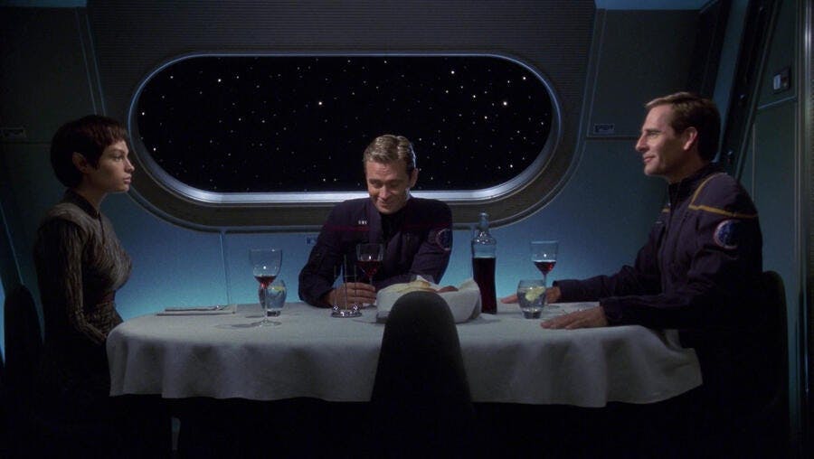 T'Pol, Trip, and Archer sit at a dinner table together. Trip and Archer are both smiling, but T'Pol has a neutral expression in 'Carbon Creek'