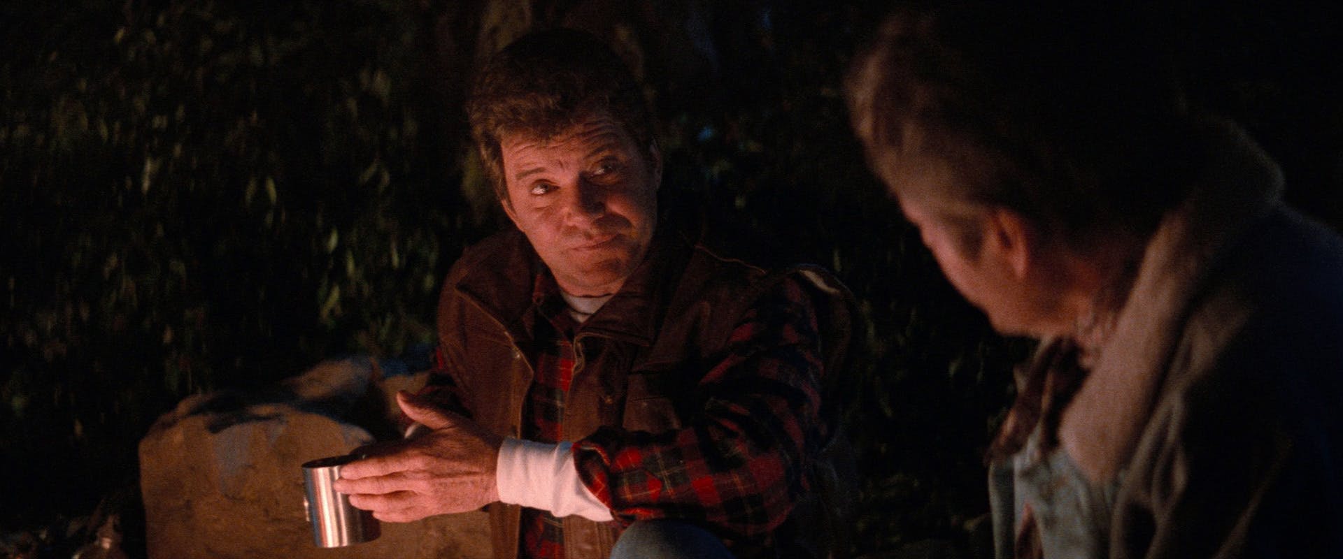 Seated around a campfire, Kirk holds his tin cup in his hand as he looks over at McCoy in Star Trek V: The Final Frontier