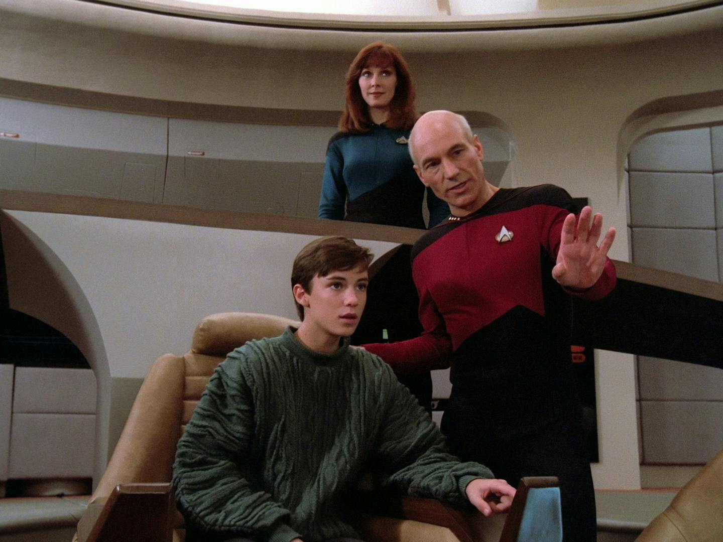 Wesley Crusher sits in the Captain's char as Jean-Luc Picard leans over and points to something in front of them as Dr. Beverly Crusher stands behind and observes in 'Encounter at Farpoint'