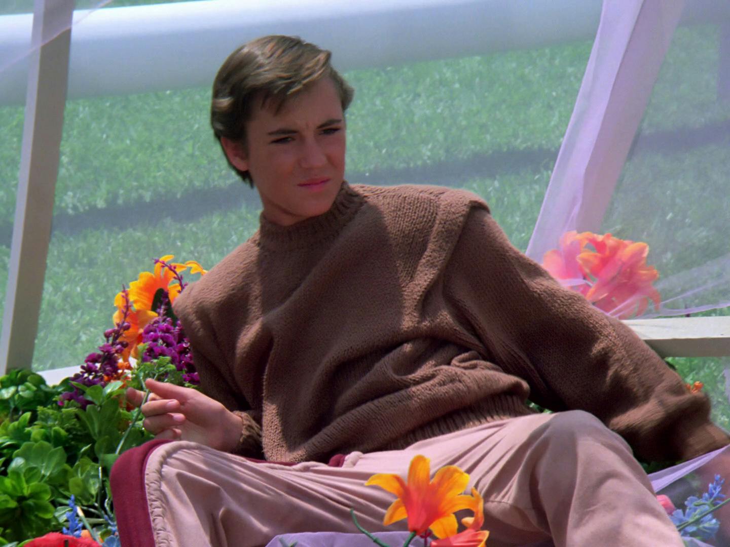 Wesley Crusher, wearing a brown knit sweater, lounges amongst the flowers on a planet's surface in 'Justice'