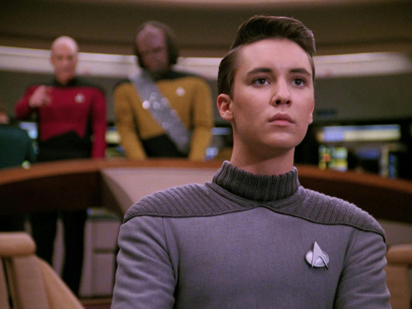 At his station on the Enterprise bridge, Wesley Crusher looks intently ahead as Picard and Worf stands behind him in 'Ménage à Troi'