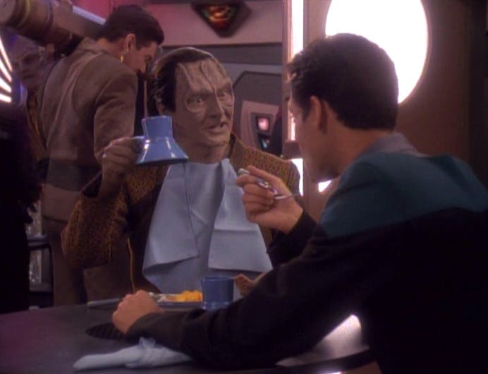 Bashir and Garak are seated at the replimat for a meal with a napkin bib in Garak's collar as he lifts his raktajino mug in 'Distant Voices'