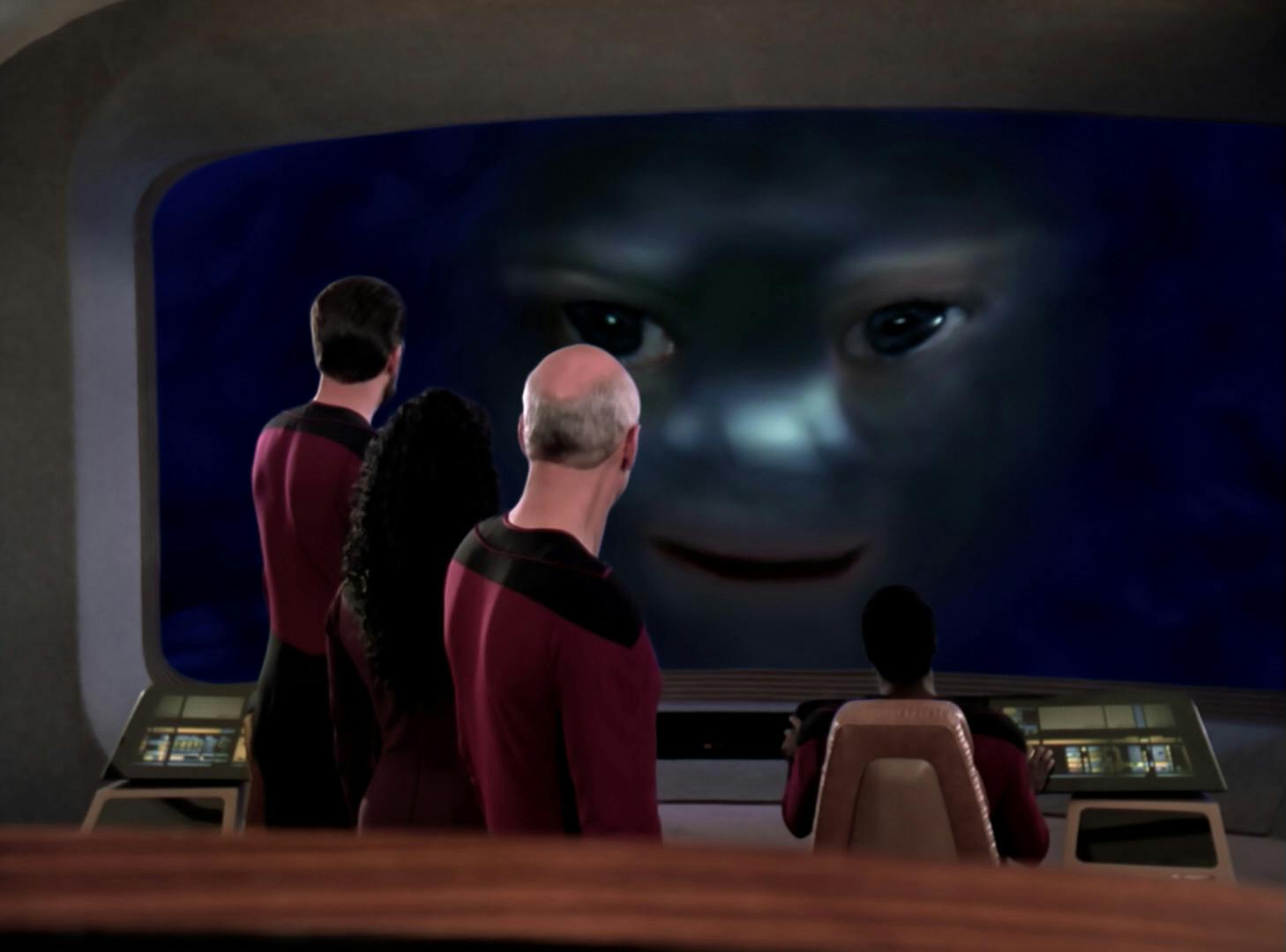 Standing on the bridge of the Enterprise-D, Will Riker, Deanna Troi, and Jean-Luc Picard look at a giant humanoid face that appears on their viewscreen in 'Where Silence Has Lease'