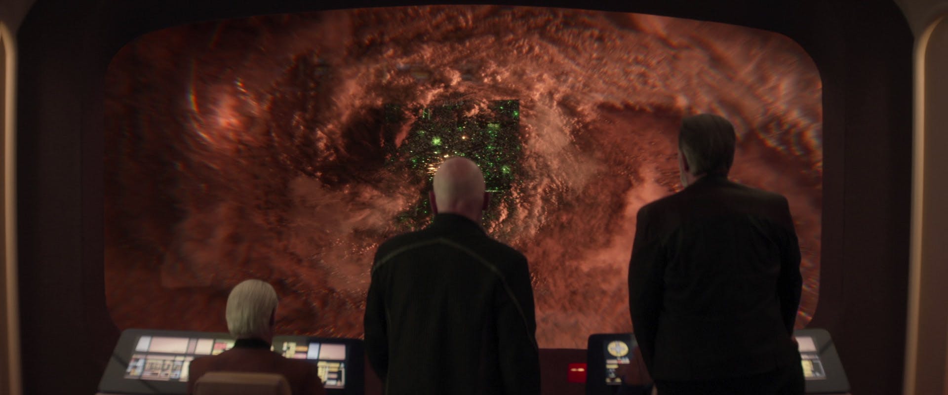 On the bridge of the Enterprise-D, a seated Data and standing Picard and Riker look out at a Borg Cube emerging from the gases surrounding Jupiter in 'The Last Generation'