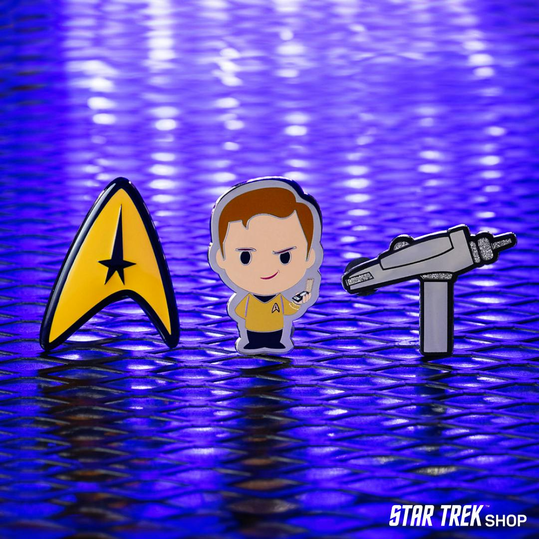 Three enamel pins (L-to-R: Yellow Delta, illustrated Kirk, & a illustrated TOS phaser) in a row on a purple background.