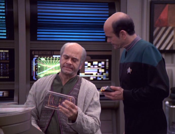 The Doctor, holding his medical tricorder, hovers over a seated Dr. Zimmerman as he looks over a chart in 'Life Line'