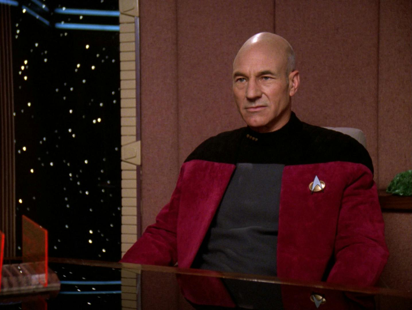 Captain Jean-Luc Picard sits in his Ready Room with his jacket uniform unzipped looking ahead at Dr. Farallon and Data in 'The Quality of Life'