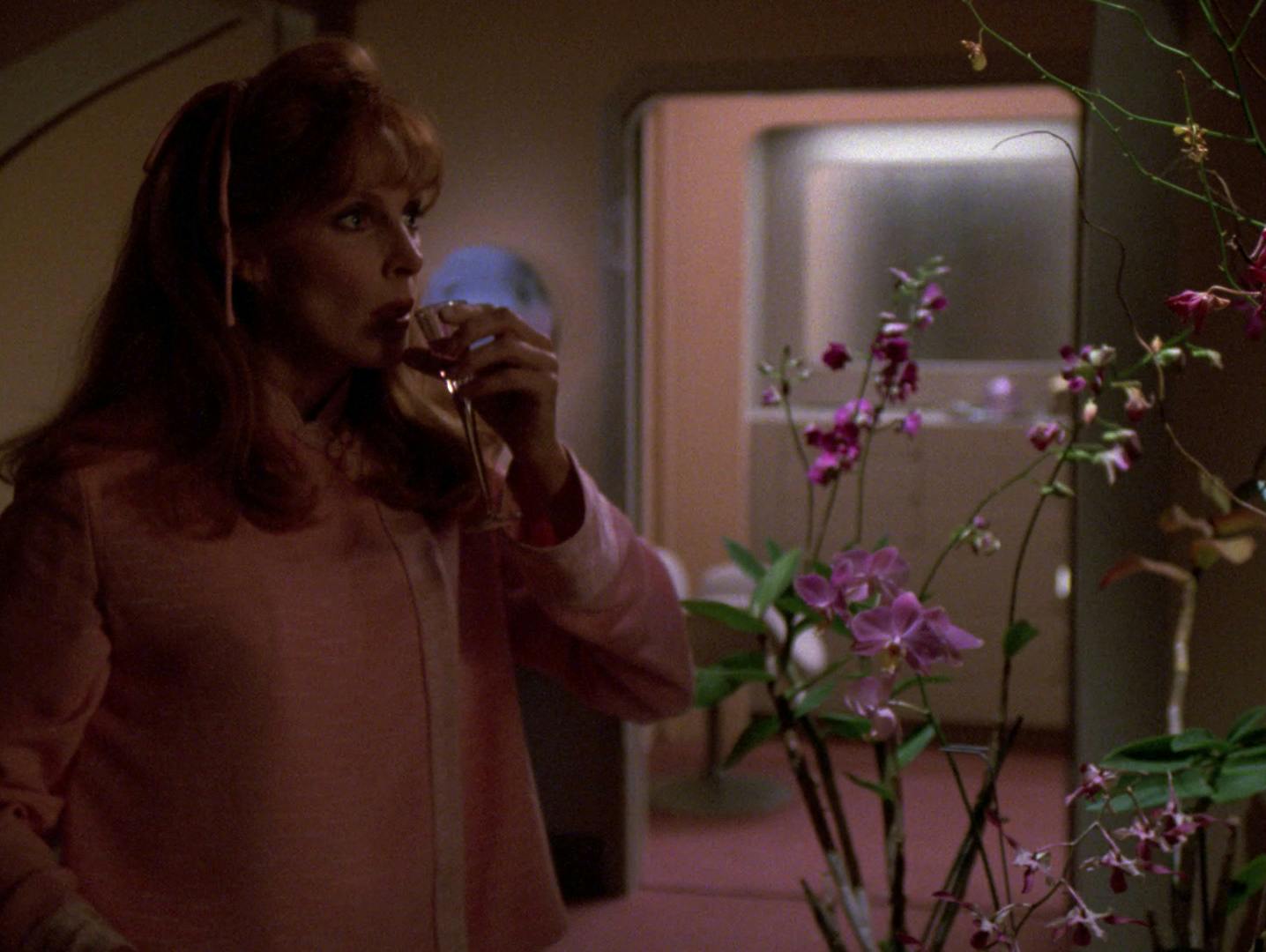 In her quarters, Beverly Crusher in her pjs drinks a night cap next to a table full of orchids in 'Cause and Effect'