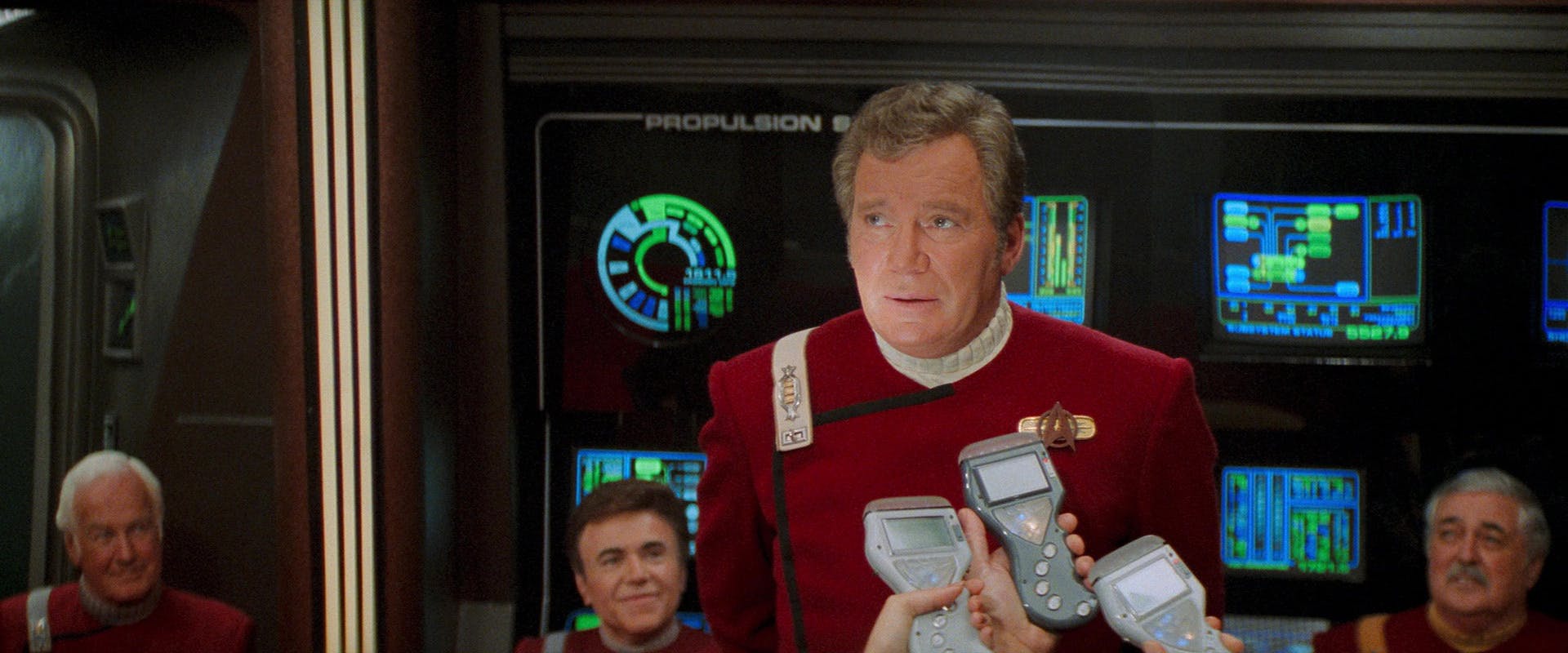 Media hold recorders in front of Captain James T. Kirk asking about the Enterprise-B and his retirement as Chekov and Scotty sit smiling behind him in 'Star Trek Generations'