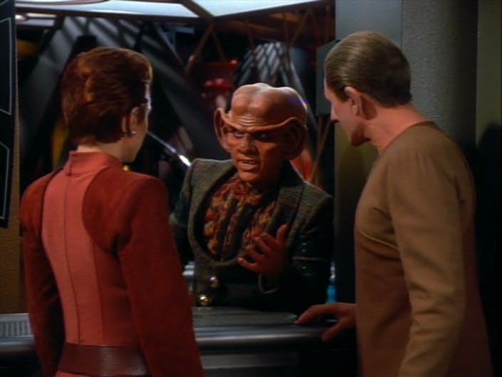 Leaning over the counter at his bar, Quark converses with Kira and Odo in 'The Collaborator'