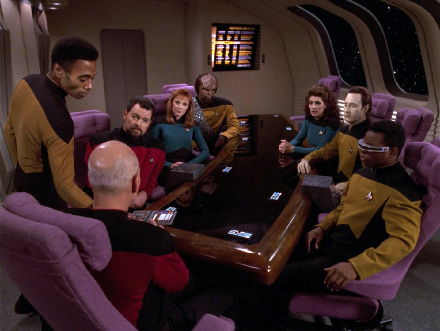 In the Observation Lounge, Ensign Armstrong hands Captain Picard a PADD as the senior staff are seated around the conference table in 'Force of Nature'