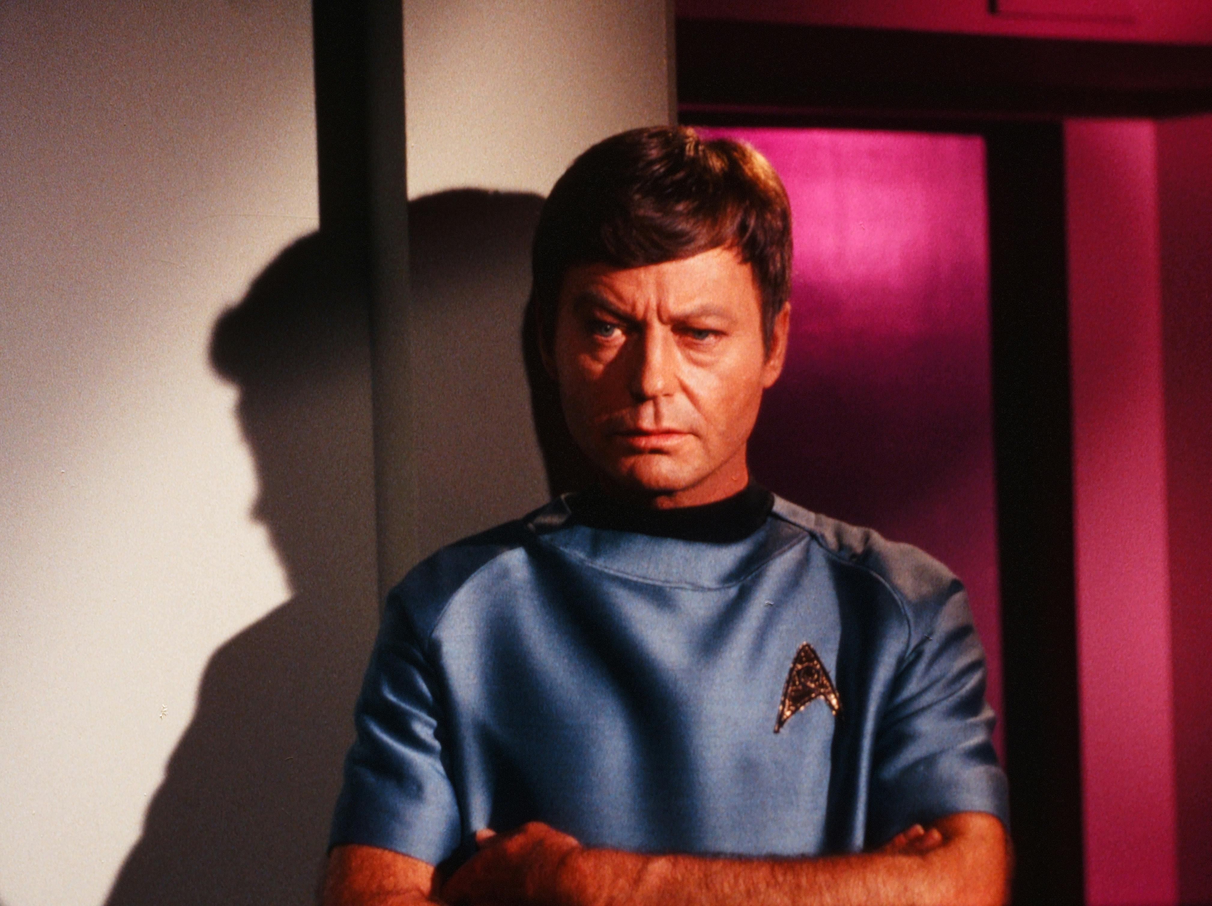 Leonard McCoy stands with his arms crossed and a stern expression in 'Obsession'