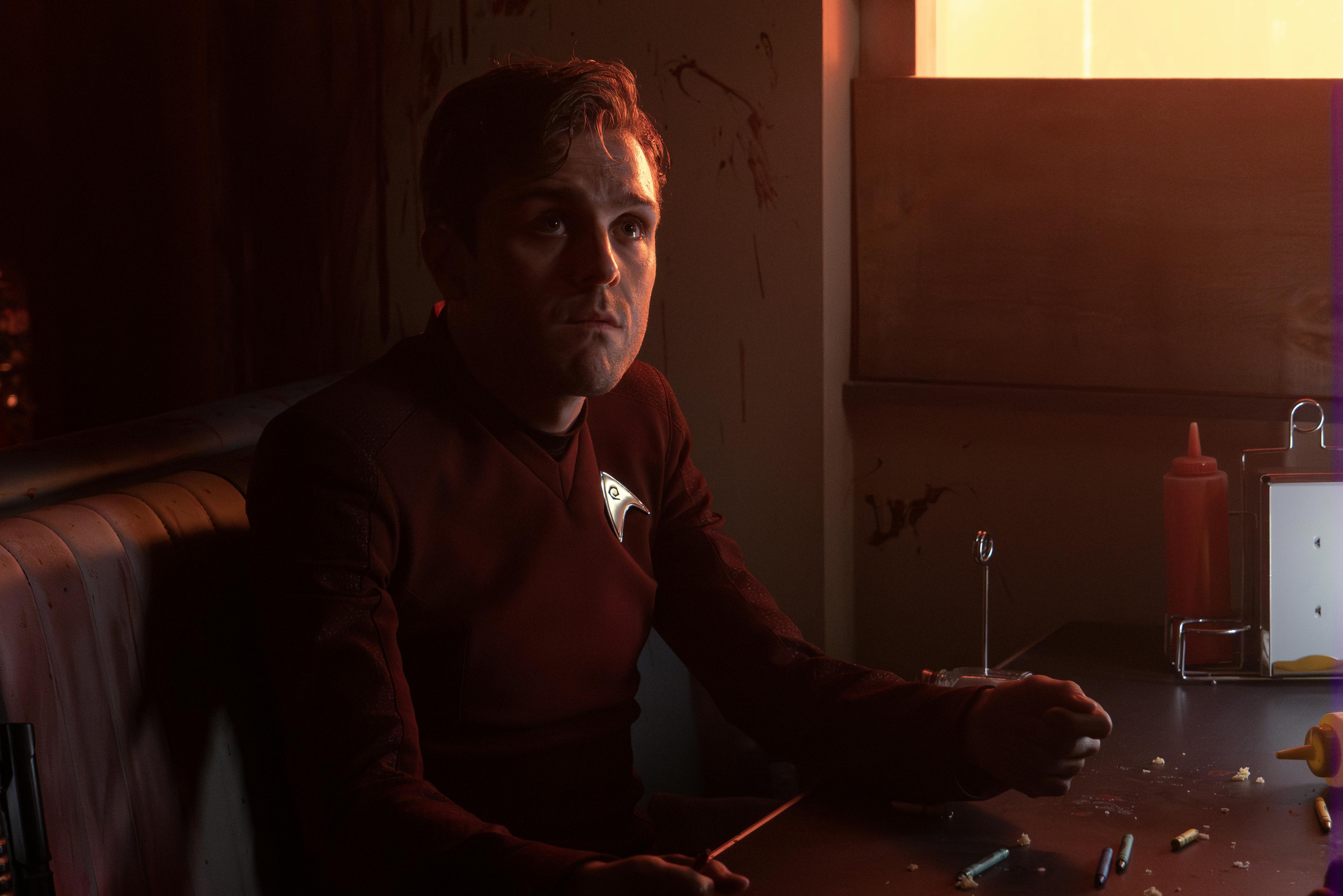 Lt. Montgomery Scott sits a diner booth and looks up towards other Starfleet officers on Parnassus Beta in 'Hegemony'