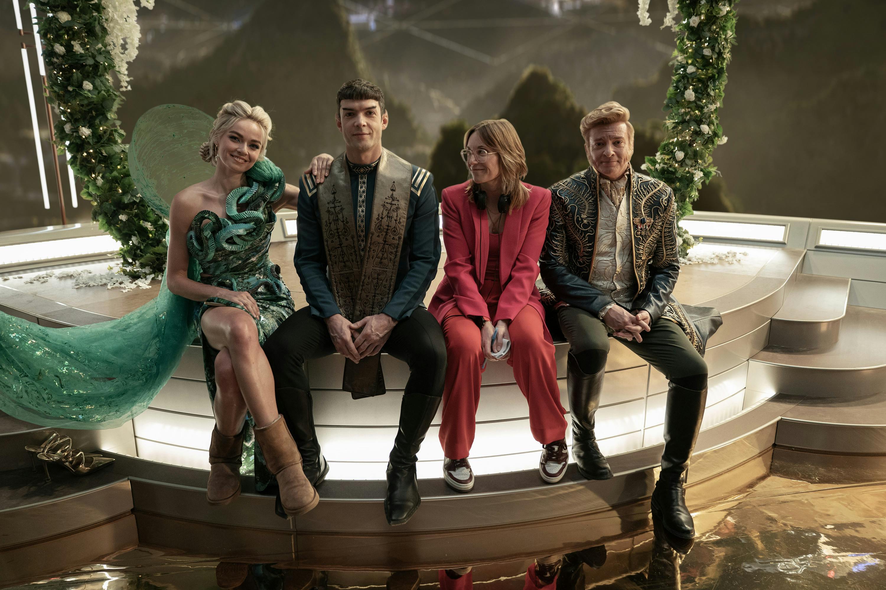 Behind-the-scenes on set of 'Wedding Bell Blues,' Jess Bush, Ethan Peck, director Jordan Canning, and Rhys Darby sit on the platform
