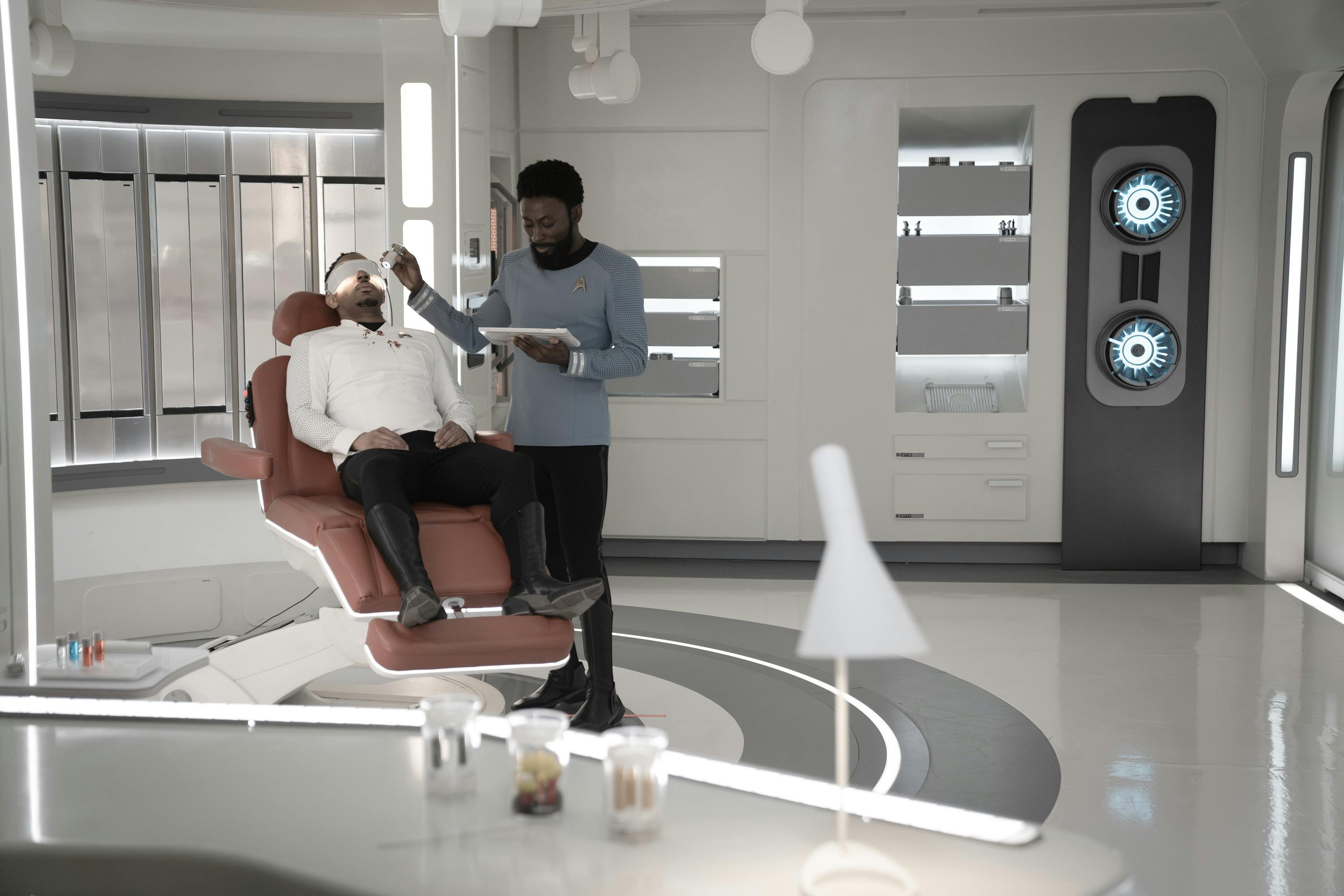 An injured Ensign Gamble lays on a biobed in Sickbay after his eyes are damaged while M'Benga runs tests in 'Through the Lens of Time'