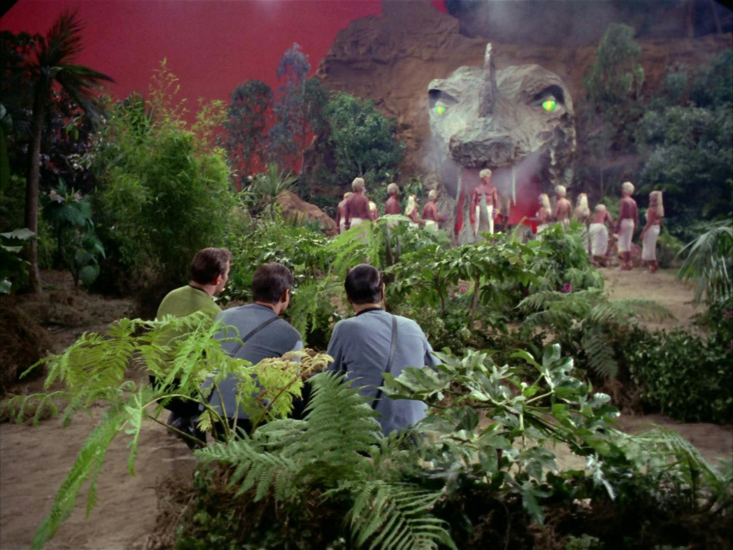 Kirk, McCoy, and Spock crouch behind some shrubbery as the people of Gamma Trianguli VI convene in front of the computer Vaal in 'The Apple'