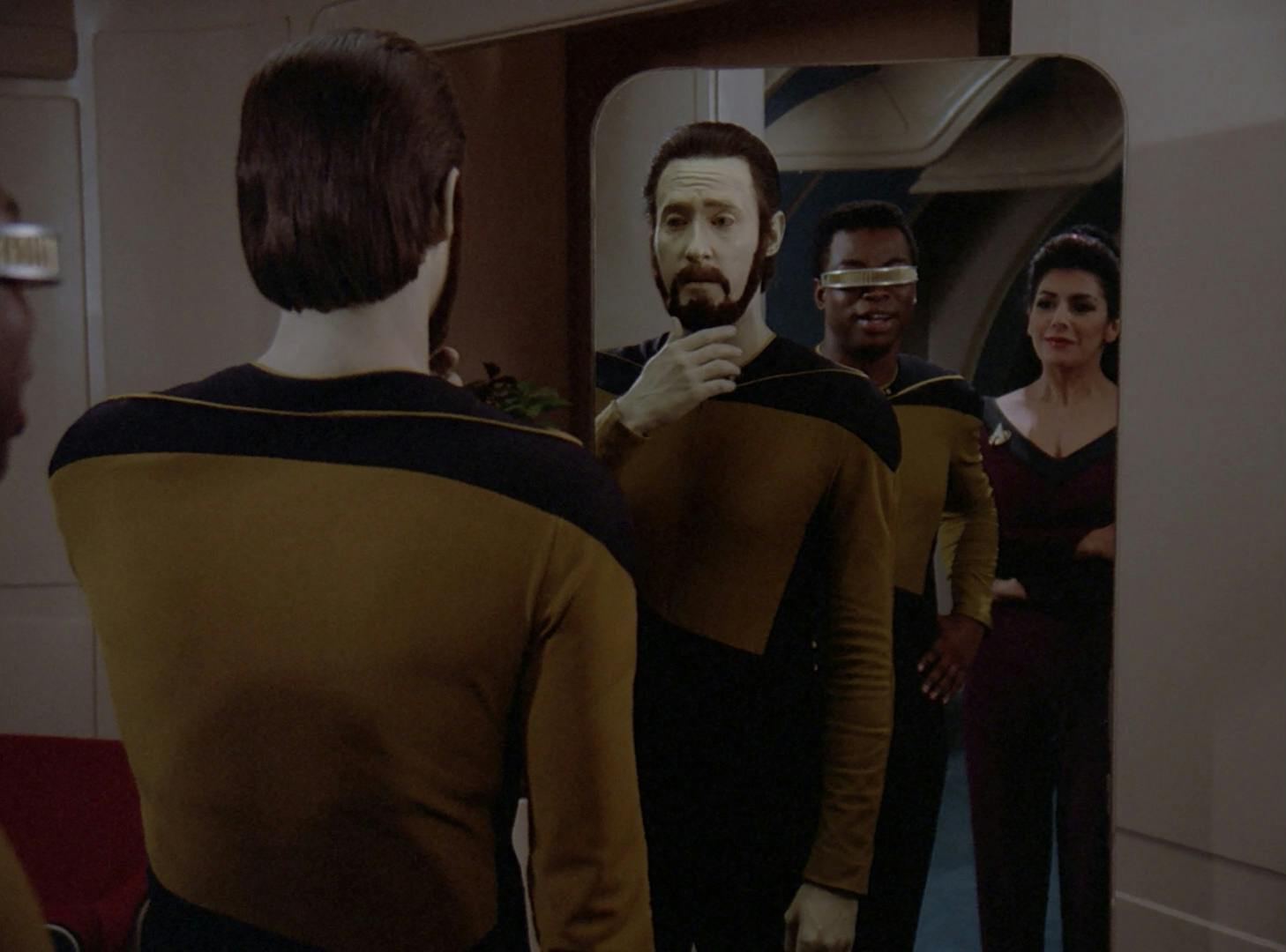 Data with a beard looks at his reflection in the mirror as Geordi La Forge and Deanna Troi amused stand behind him in 'The Schizoid Man'