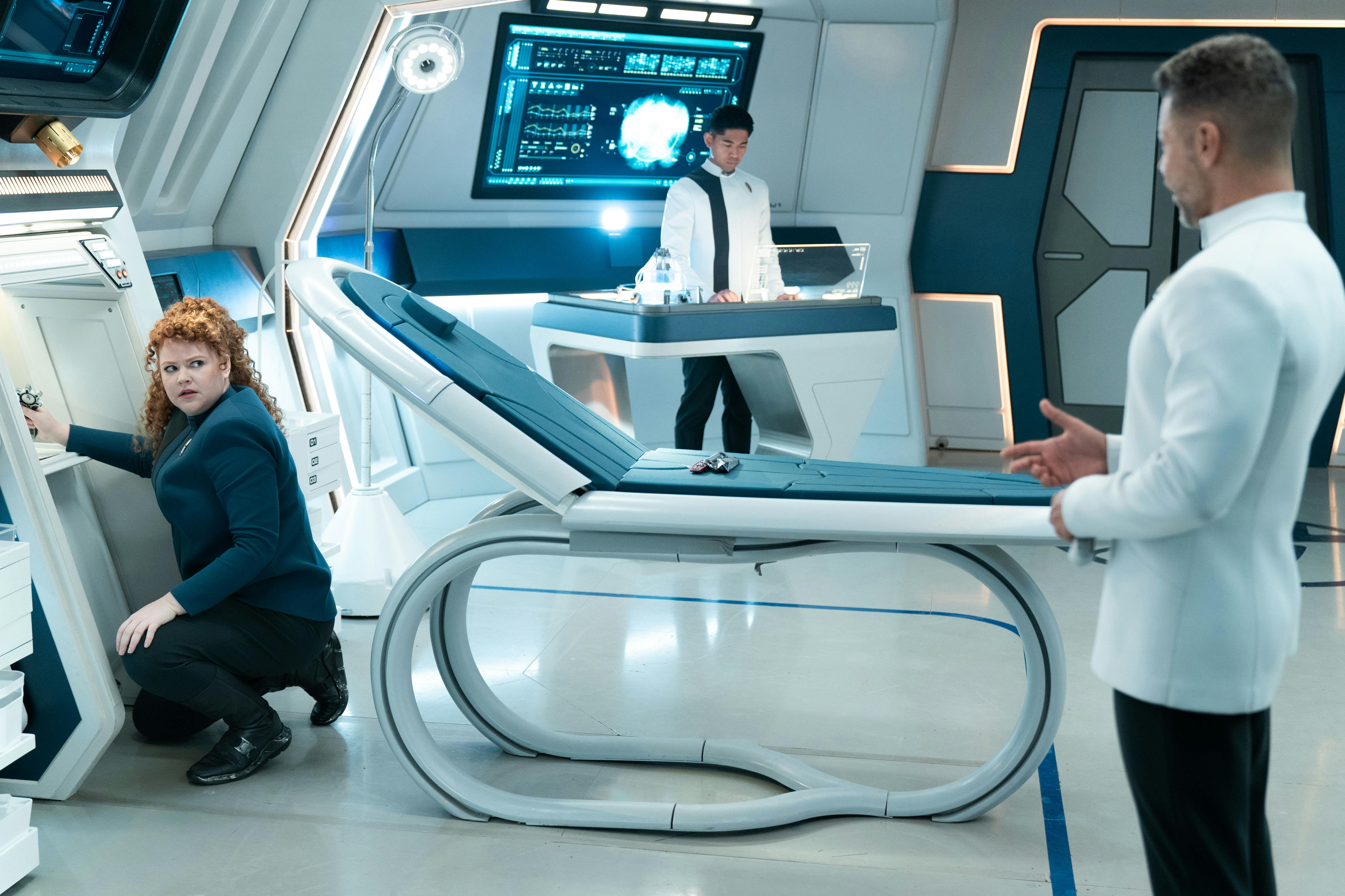 In Sickbay, Tilly kneels down as she tracks a conduit in a panel as she looks over her shoulder at Hugh Culber in 'Mirrors'