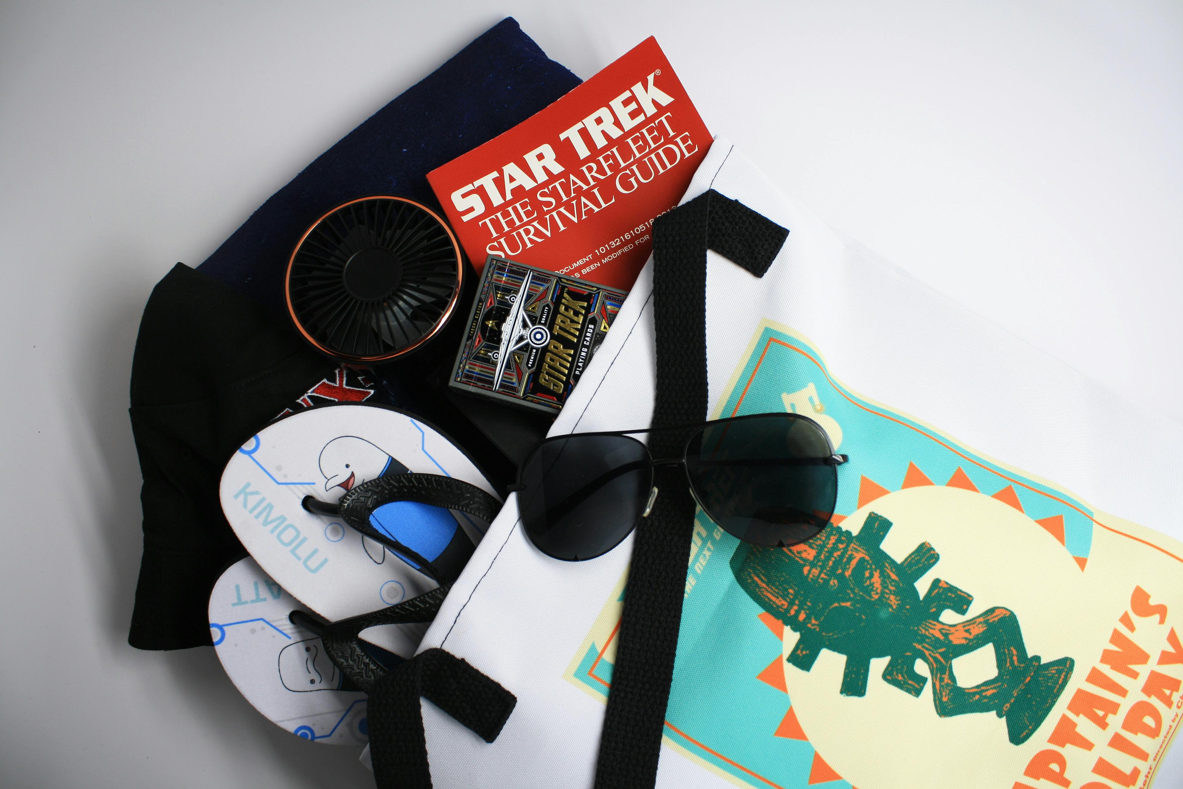A Captain's Holiday canvas tote holding a pair of Star Trek: Lower Decks flip flops, a NX-01 bucket hat, a handheld fan, Star Trek playing card, Starfleet Survival Guide, and a pair of sunglasses