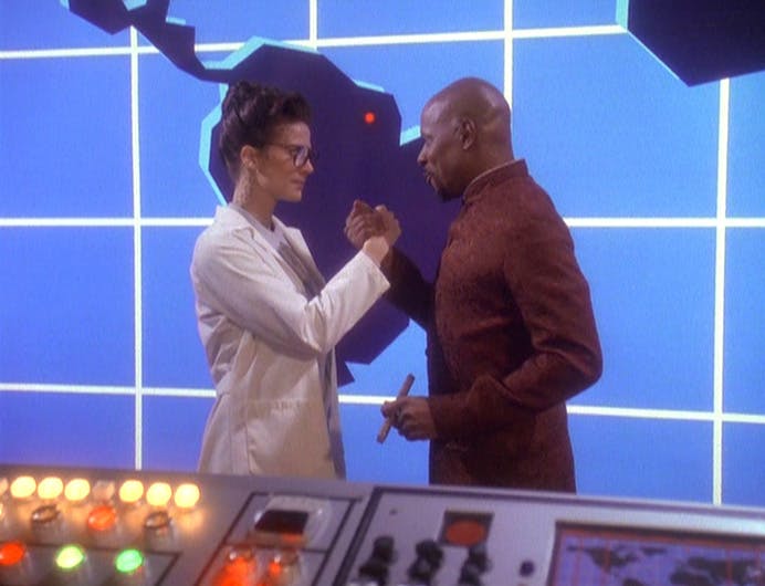 In a holoprogram, Jadzia Dax as Dr. Honey Bare and Ben Sisko as Dr. Noah clasp their hands in a pact in front of a blue viewscreen with a map in 'Our Man Bashir'