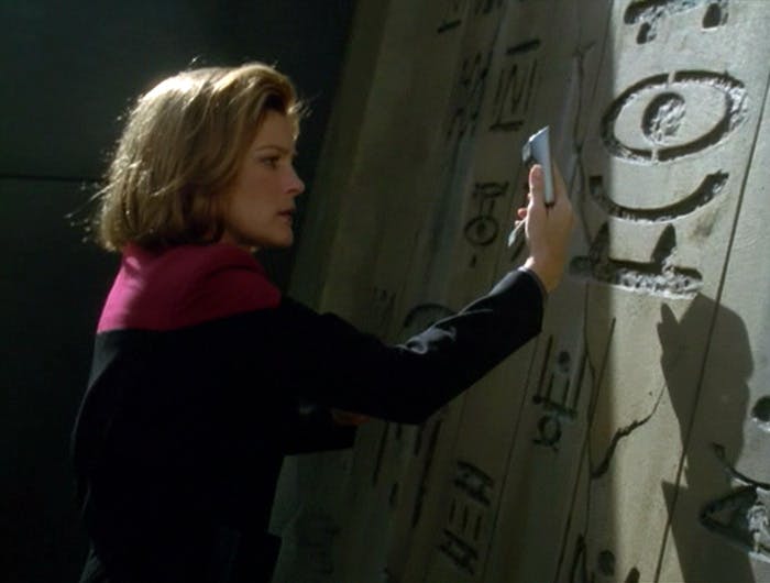 Captain Janeway holds her tricorder up to a wall with engraving on a memorial monument in 'Memorial'