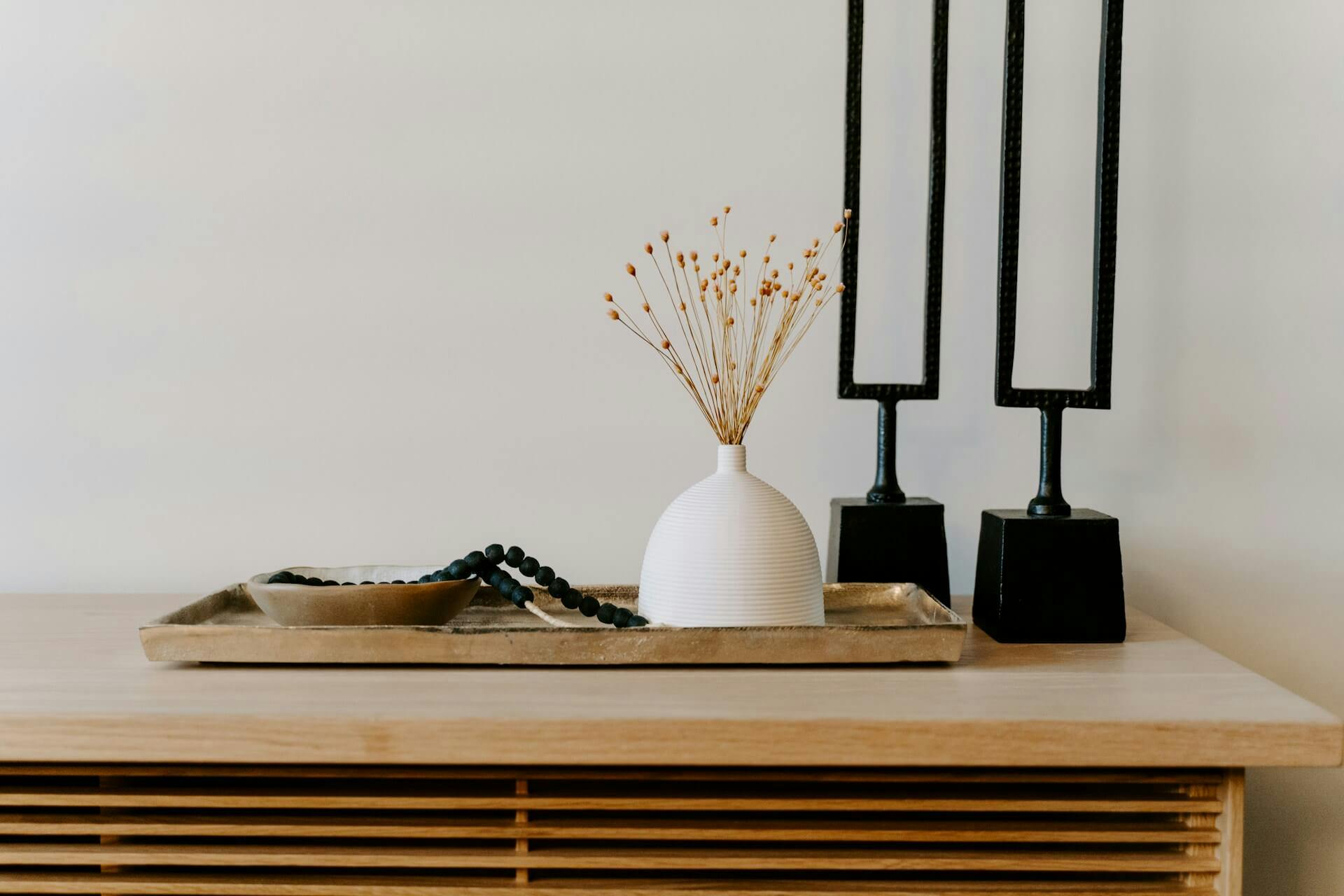 A minimalist decor arrangement on a light wooden surface, featuring a textured white ceramic vase with dried floral stems, placed on a metallic tray alongside a black bead garland, two modern, black geometric sculptures.