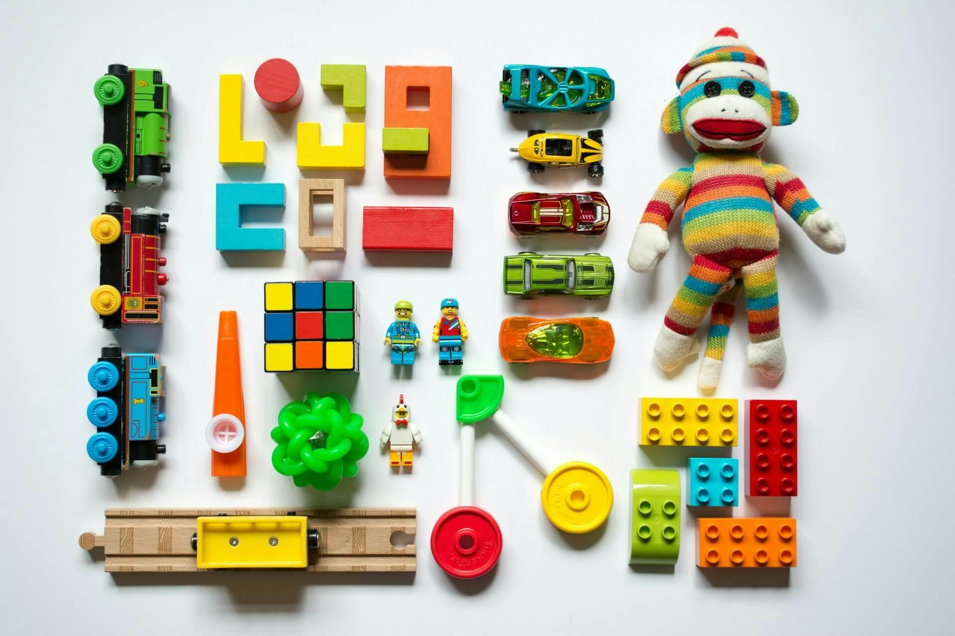 A vibrant collection of toys arranged, including colourful wooden trains, foam alphabet blocks, miniature toy cars, a Rubik’s Cube, building blocks, a striped sock monkey plush toy, a whistle, a spinning top and lego.