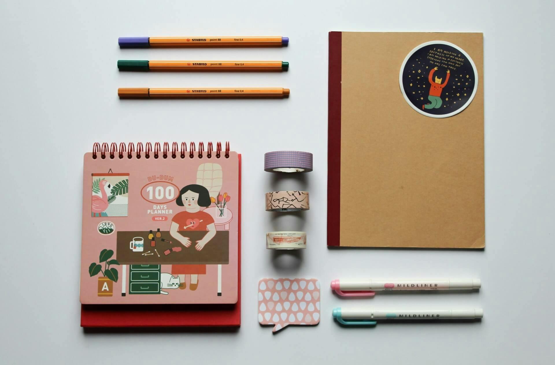 A neatly arranged stationery set featuring a spiral-bound "100 Days Planner" with a colourful illustrated cover, three vibrant fine-tip pens in orange, green, and purple, and a brown notebook with a whimsical celestial sticker. Alongside are three decorative washi tapes, two pastel highlighters, and a pink speech bubble sticky note, all laid out on a clean white surface, perfect for planning or creative journaling.