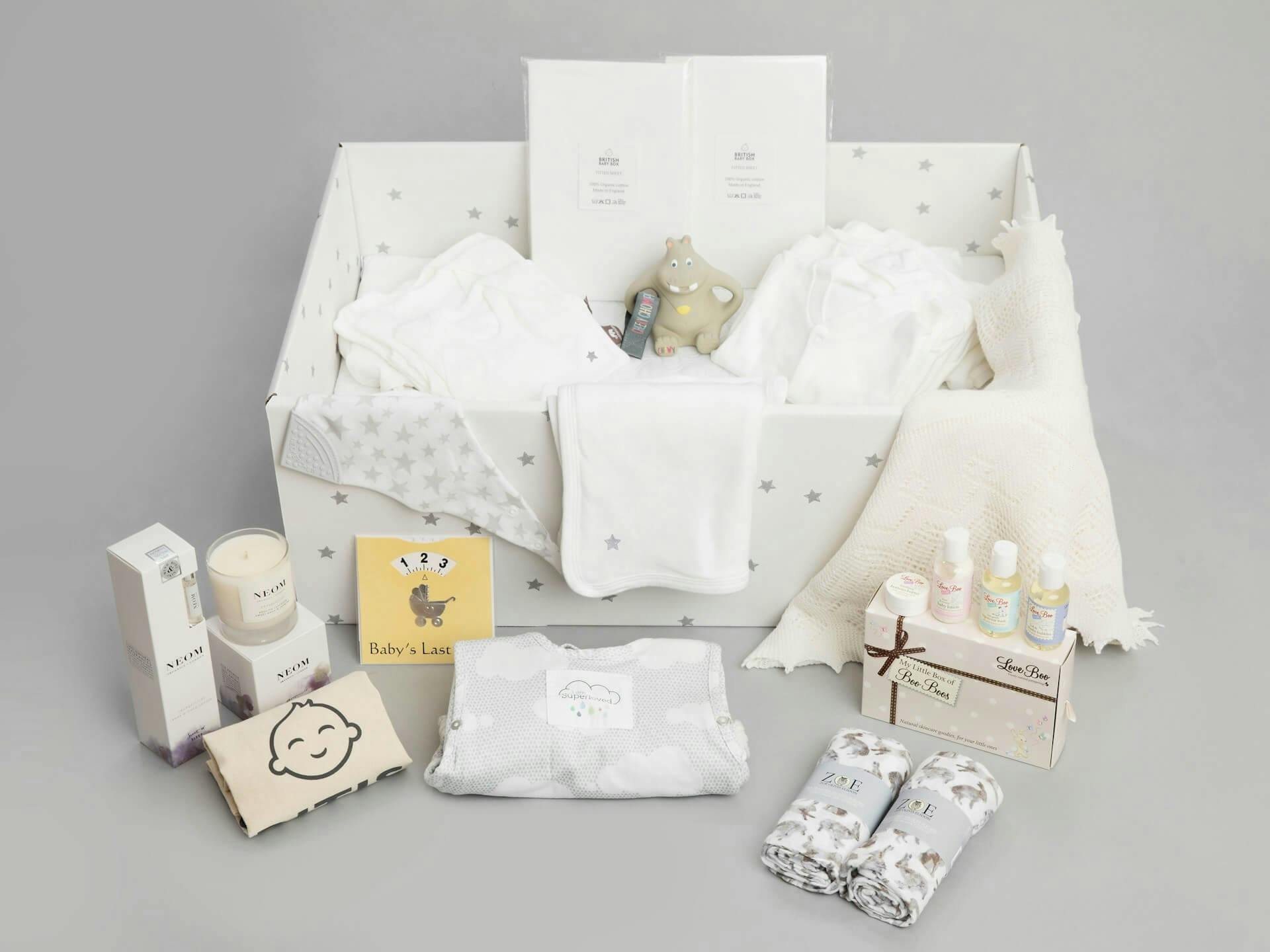 A baby gift set displayed in a star-patterned box, containing white baby clothes, blankets, a hippo-shaped teething toy, a set of skincare products, baby milestone cards, and other baby essentials like muslin cloths and comfort items.