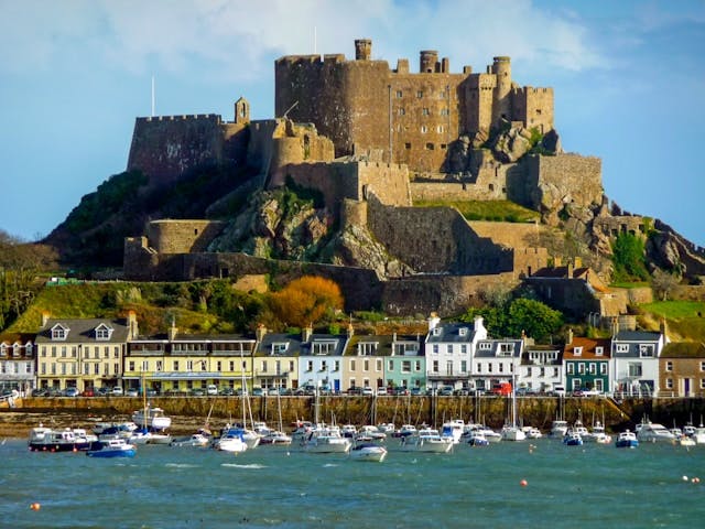 coastal town in jersey, channel islands, featuring a castle atop a hill overlooking a marina with boats.