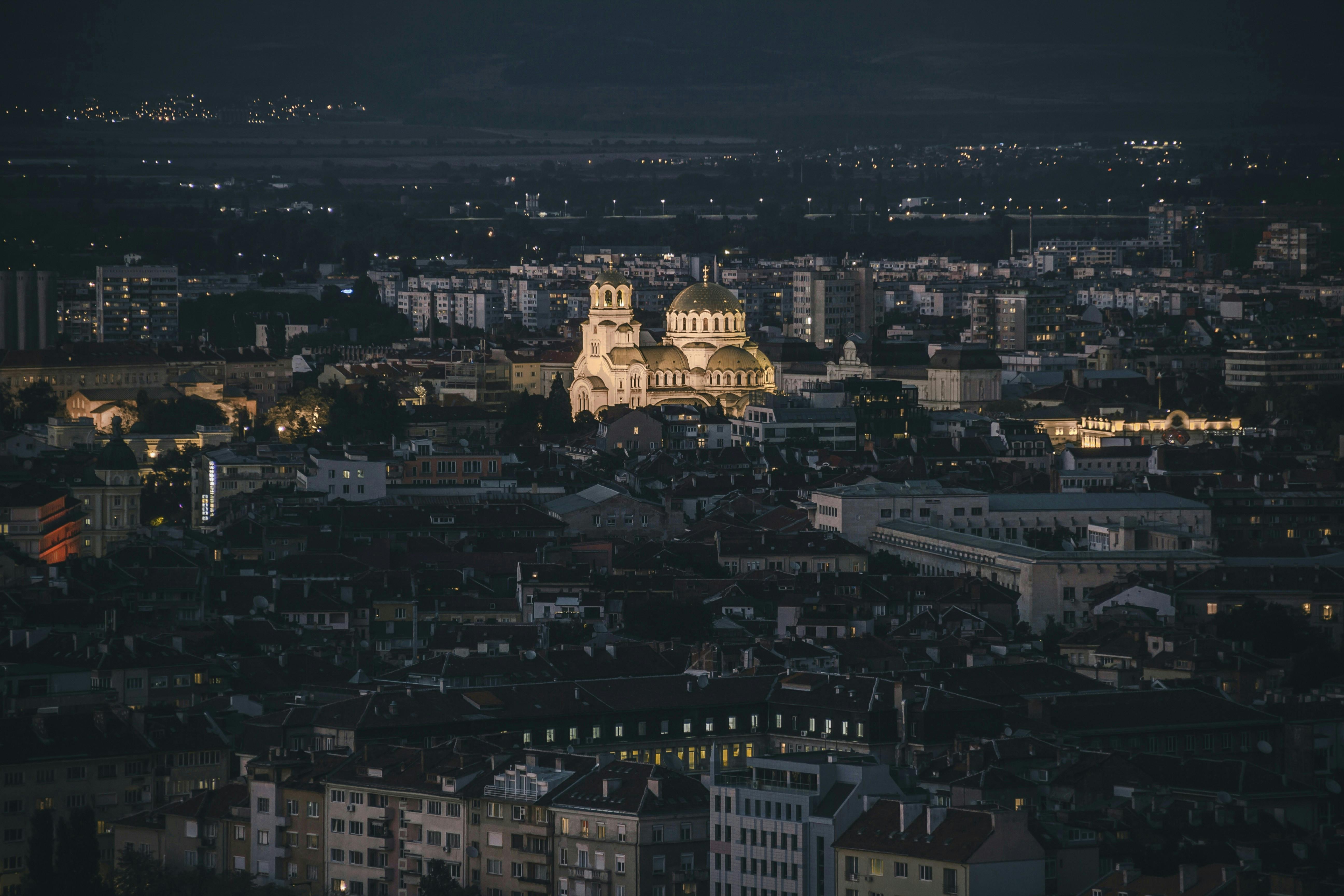 A night view of Sofia, Bulgaria, with its iconic domes standing out in the cityscape. The surrounding urban area  are buildings and streets.