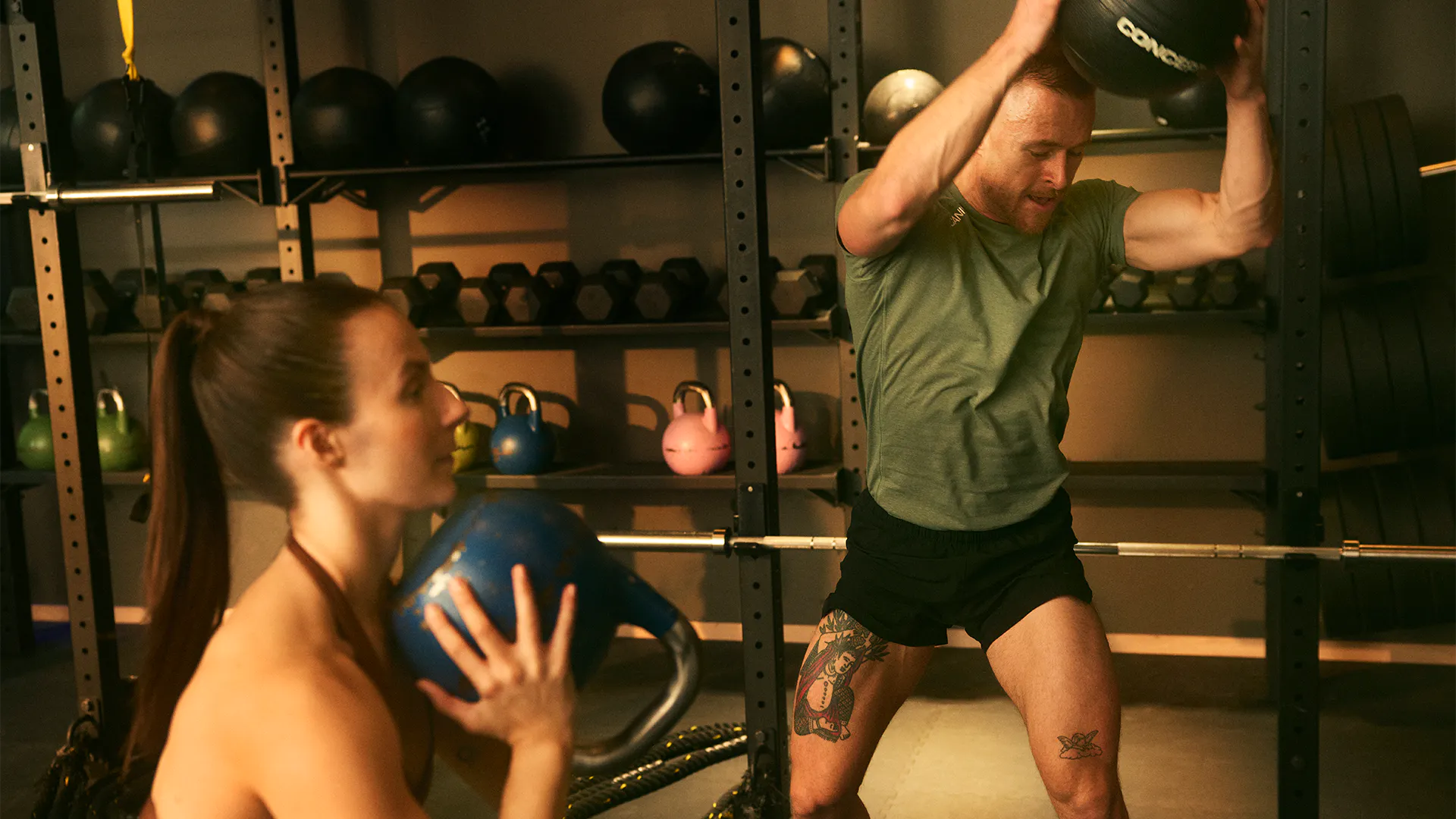 Kvinna med mörkt hår och hästsvans lyfter en kettlebell under hakan, en man i grön T-shirt och tatueringar kastar en medicinboll mot marken