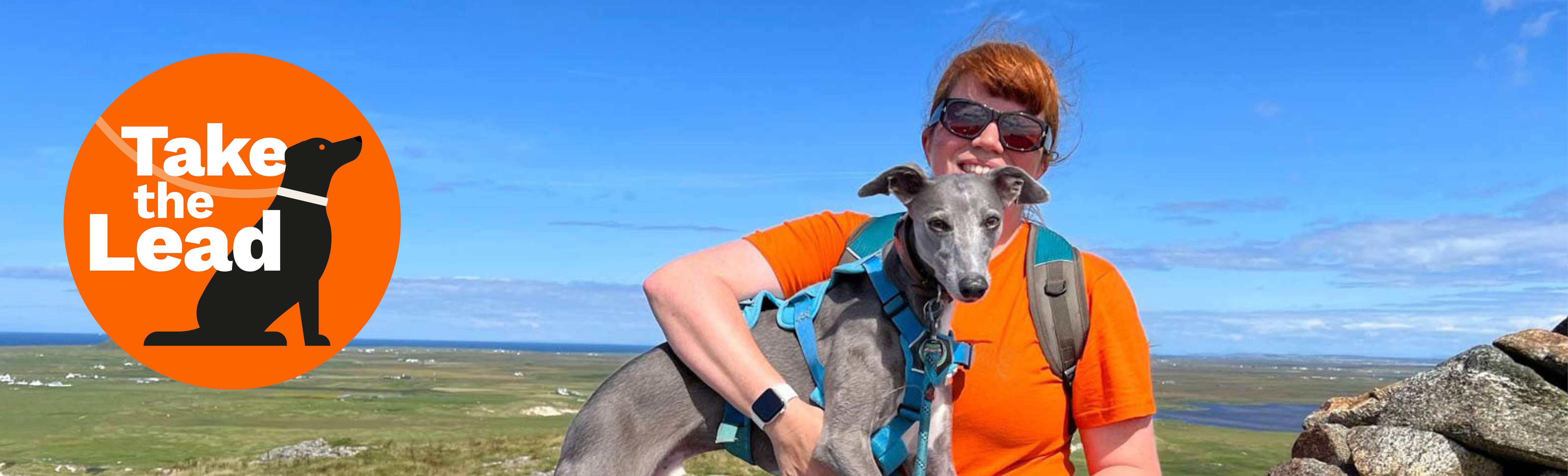 Person sitting outdoors on rocky ground holding a greyhound in a teal harness, with green fields and blue sky in the background. An orange “Take the Lead” logo with a dog silhouette is on the left.